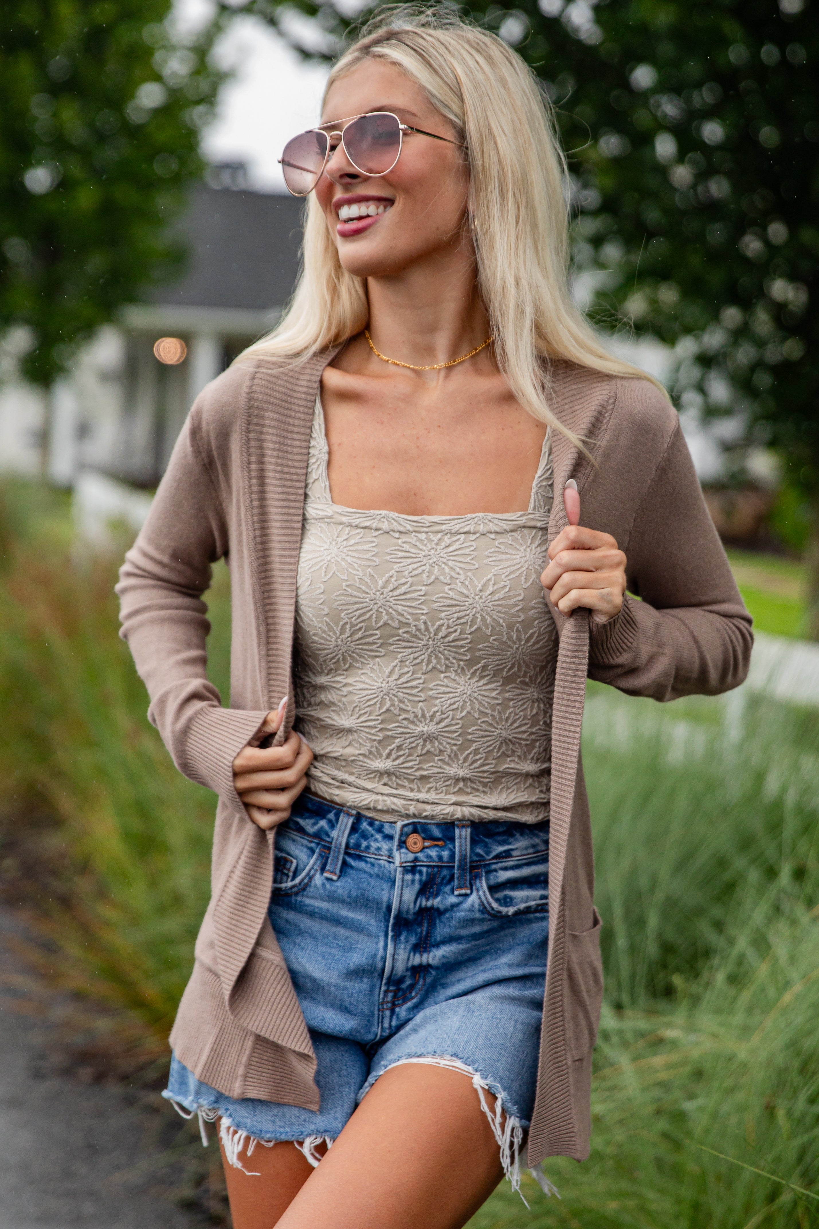 Woman wearing a beige cardigan, lace top, and denim shorts outdoors.