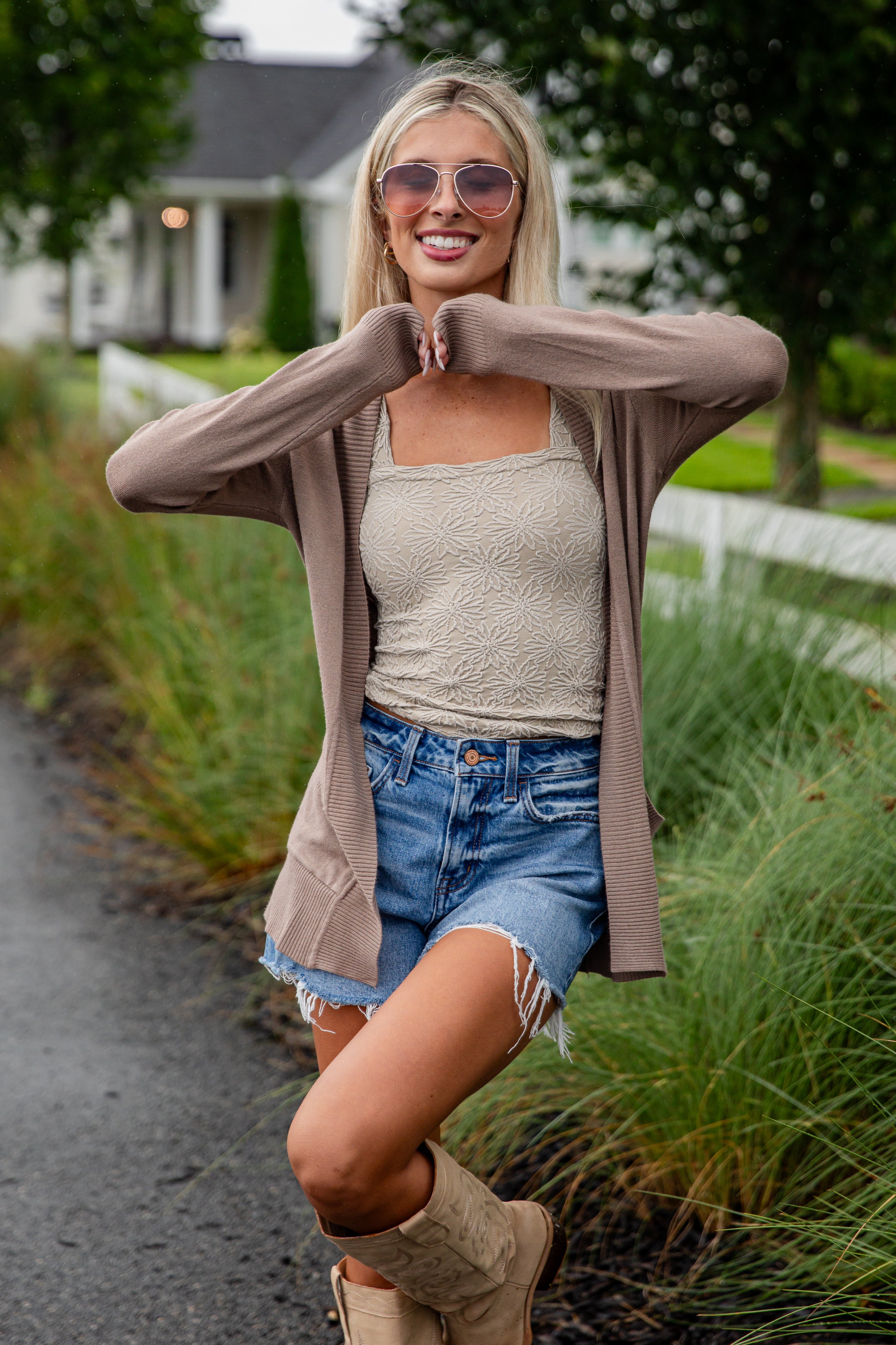 Woman wearing a beige cardigan, tank top, denim shorts, and boots outdoors.