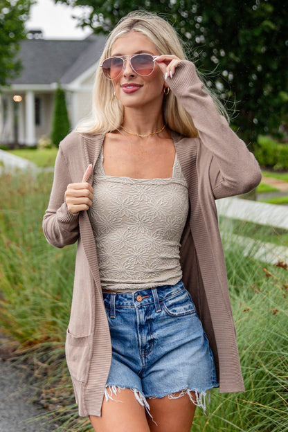 Woman walking outdoors wearing a beige cardigan, tank top, denim shorts, and knee-high boots.
