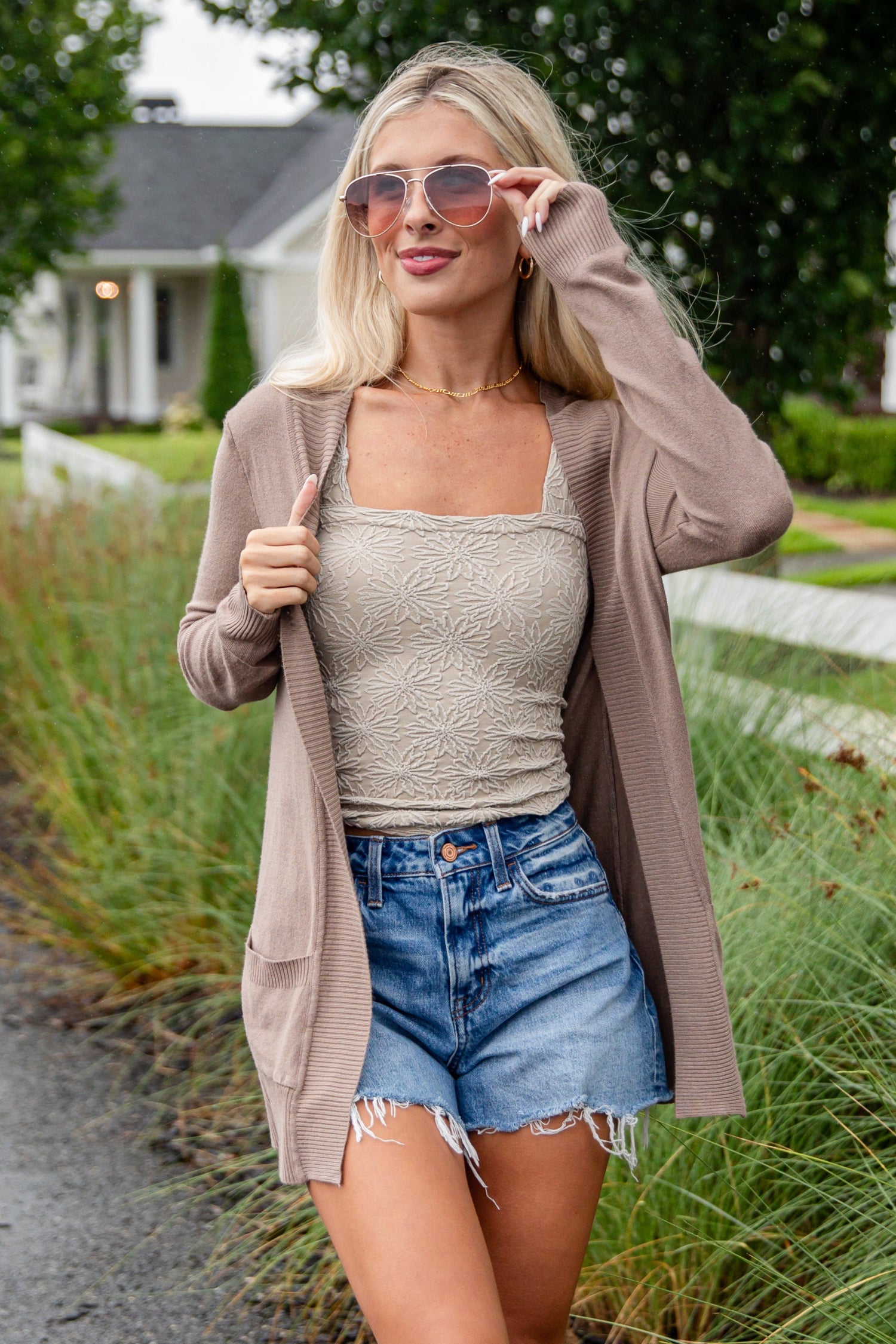 Woman walking outdoors wearing a beige cardigan, tank top, denim shorts, and knee-high boots.