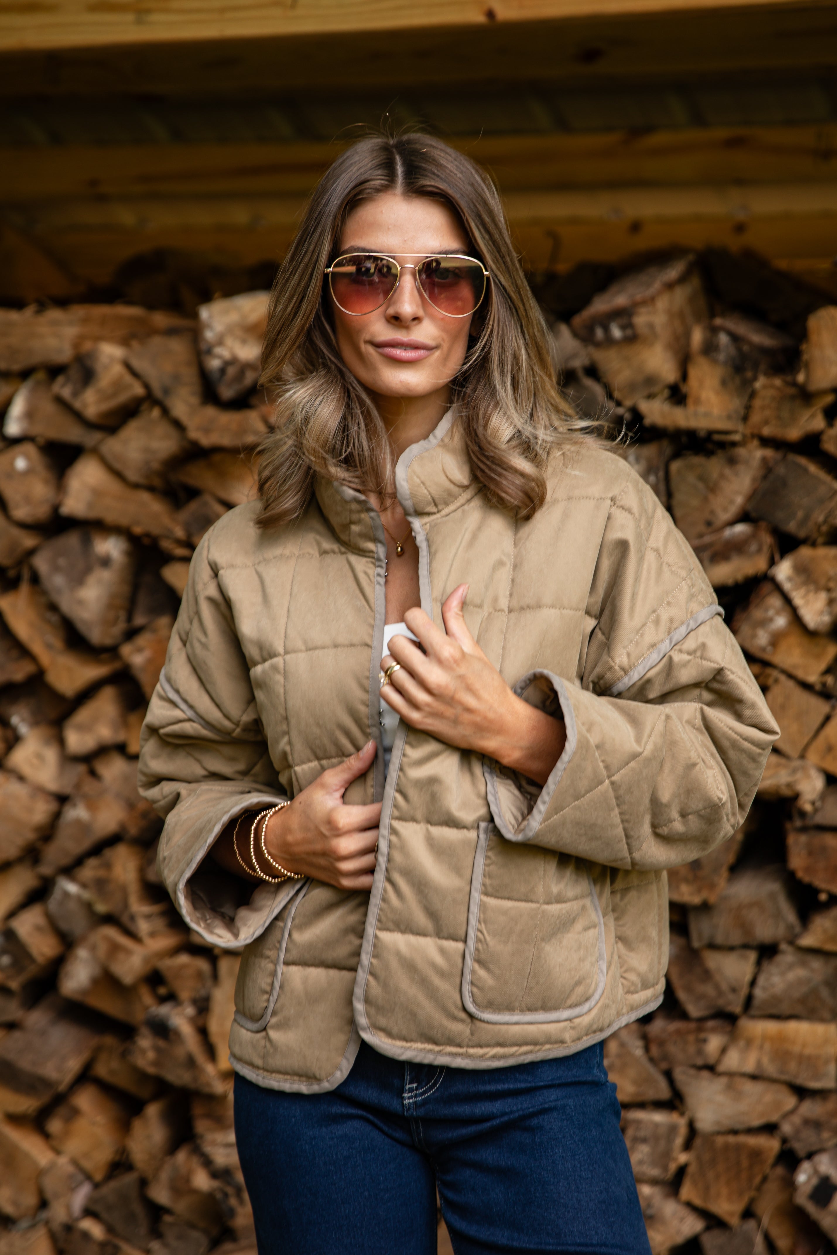Woman wearing a beige quilted jacket and sunglasses in front of stacked wood.