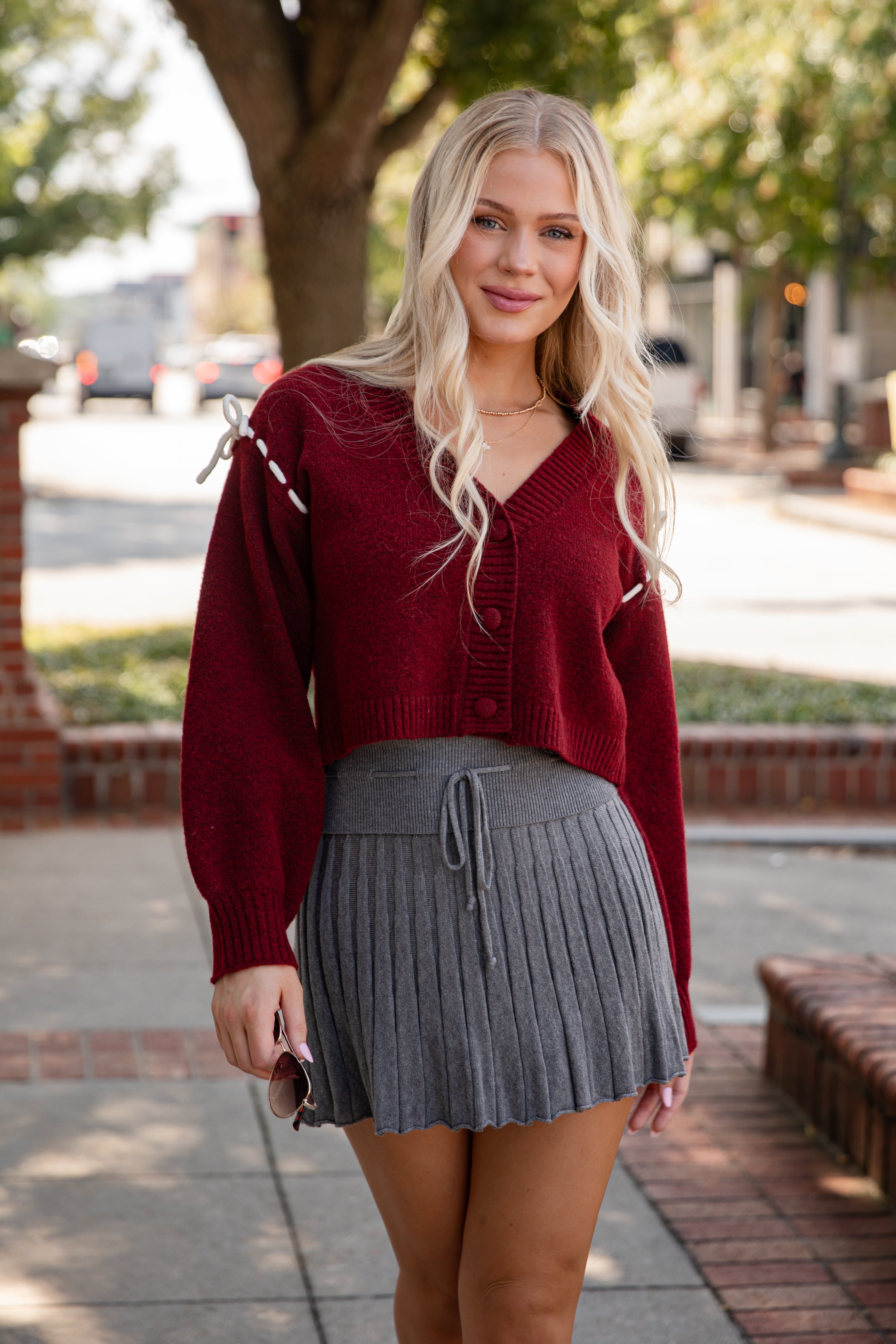 Woman wearing a red cardigan and gray pleated skirt on a sidewalk.