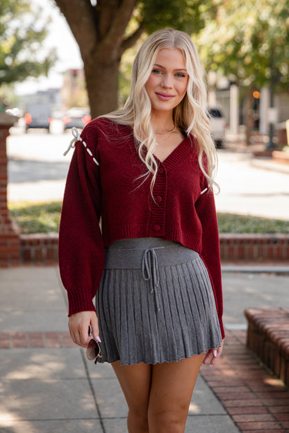Woman wearing a red cardigan and gray pleated skirt on a sidewalk.