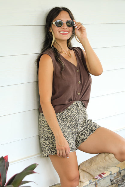 Woman in brown sleeveless top and patterned shorts standing against a white wall with plants around.