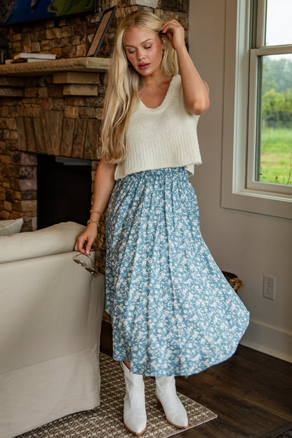Woman in a living room wearing a white top and blue floral skirt.