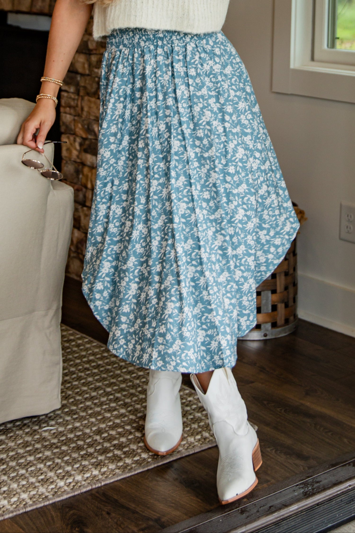 Woman in a white sleeveless top and blue floral skirt standing in a room with a fireplace and window.