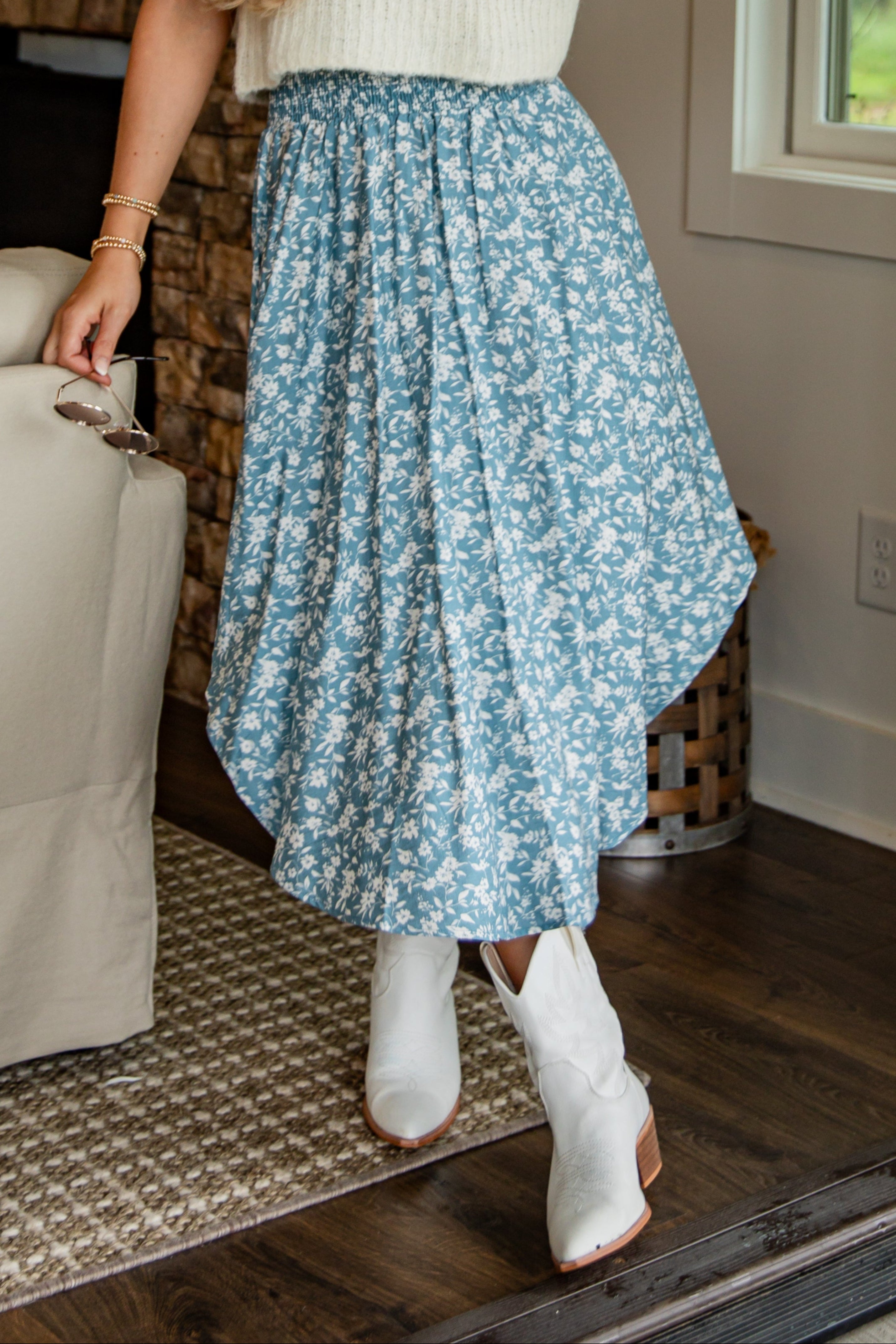 Woman in a white sleeveless top and blue floral skirt standing in a room with a fireplace and window.