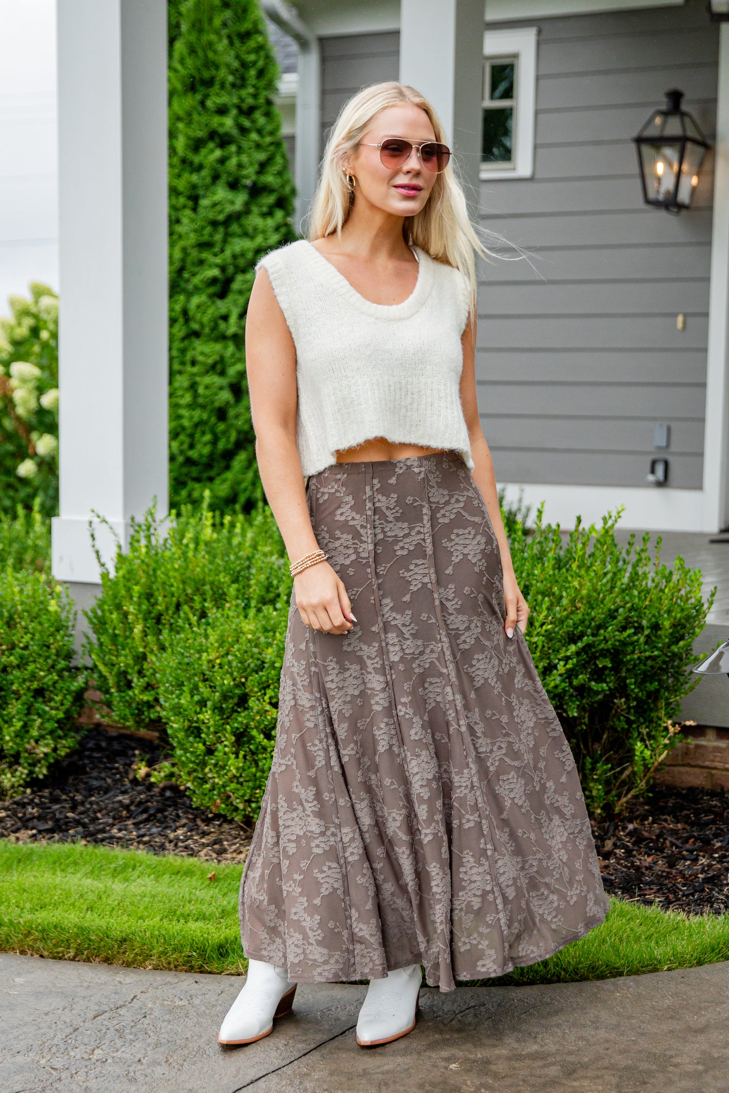 Woman wearing a white sleeveless top and brown patterned skirt standing outside a house.