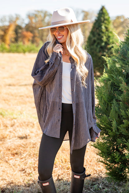 Woman standing outdoors in a field with trees, wearing a plaid shirt, white top, black pants, and boots.