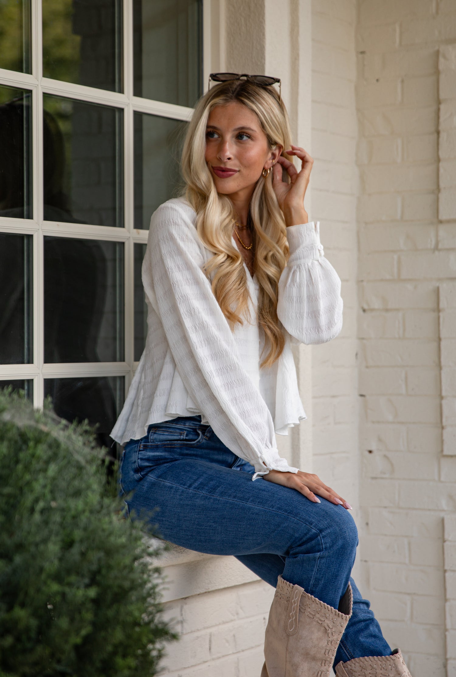 Woman in a white blouse and blue jeans sitting on a ledge outdoors.