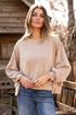 Woman wearing a beige sweater and blue jeans standing outdoors near a wooden cabin.