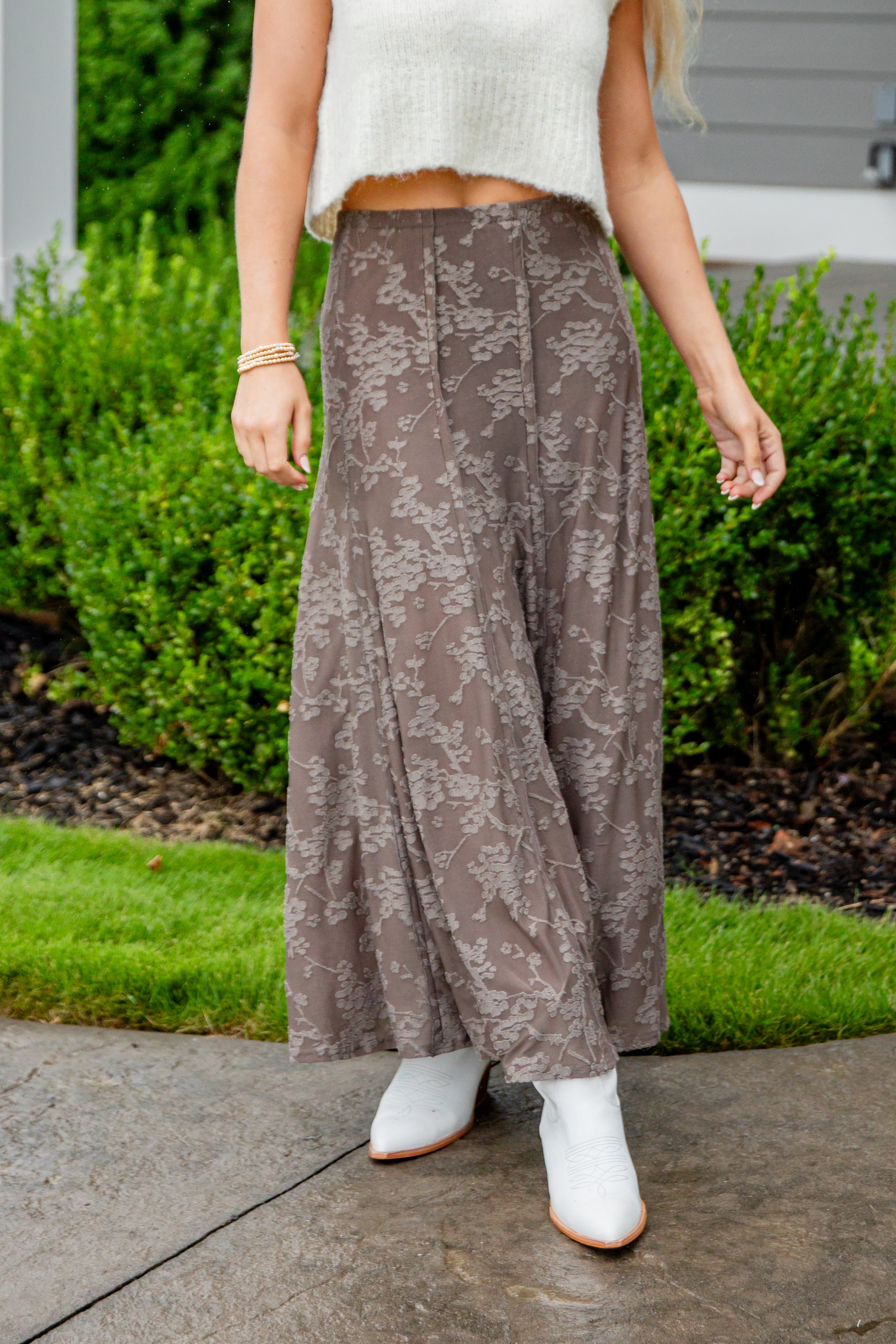 Person wearing a patterned skirt and white shoes on a sidewalk with greenery in the background