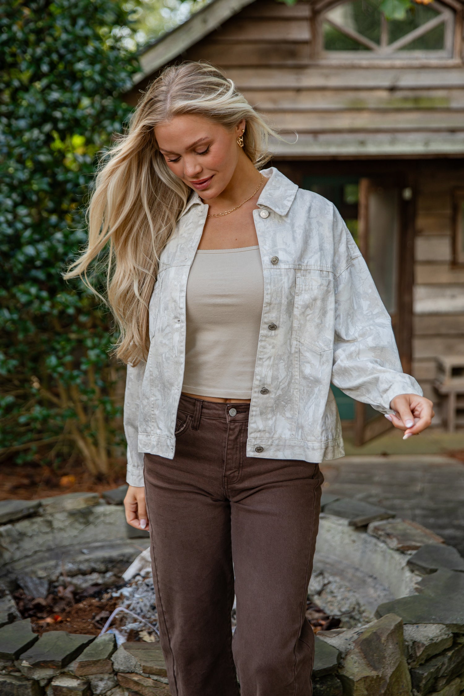 Woman wearing a light jacket over a beige top and brown pants outdoors.