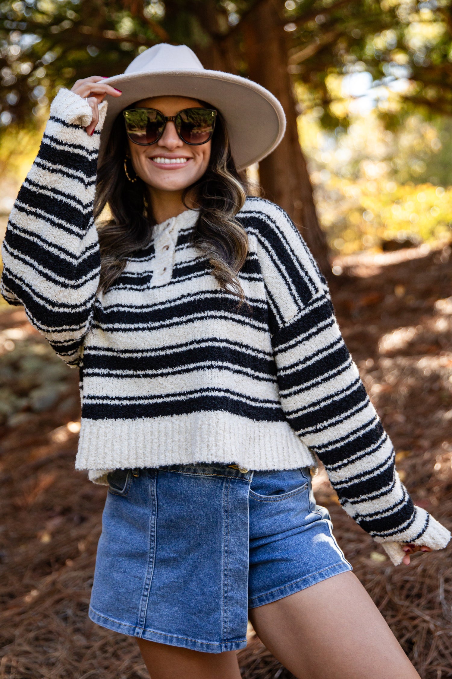 Woman wearing a striped sweater, denim shorts, and a wide-brimmed hat outdoors.