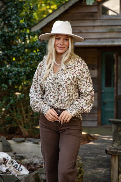 Woman wearing a patterned blouse and brown pants with a white hat outdoors.