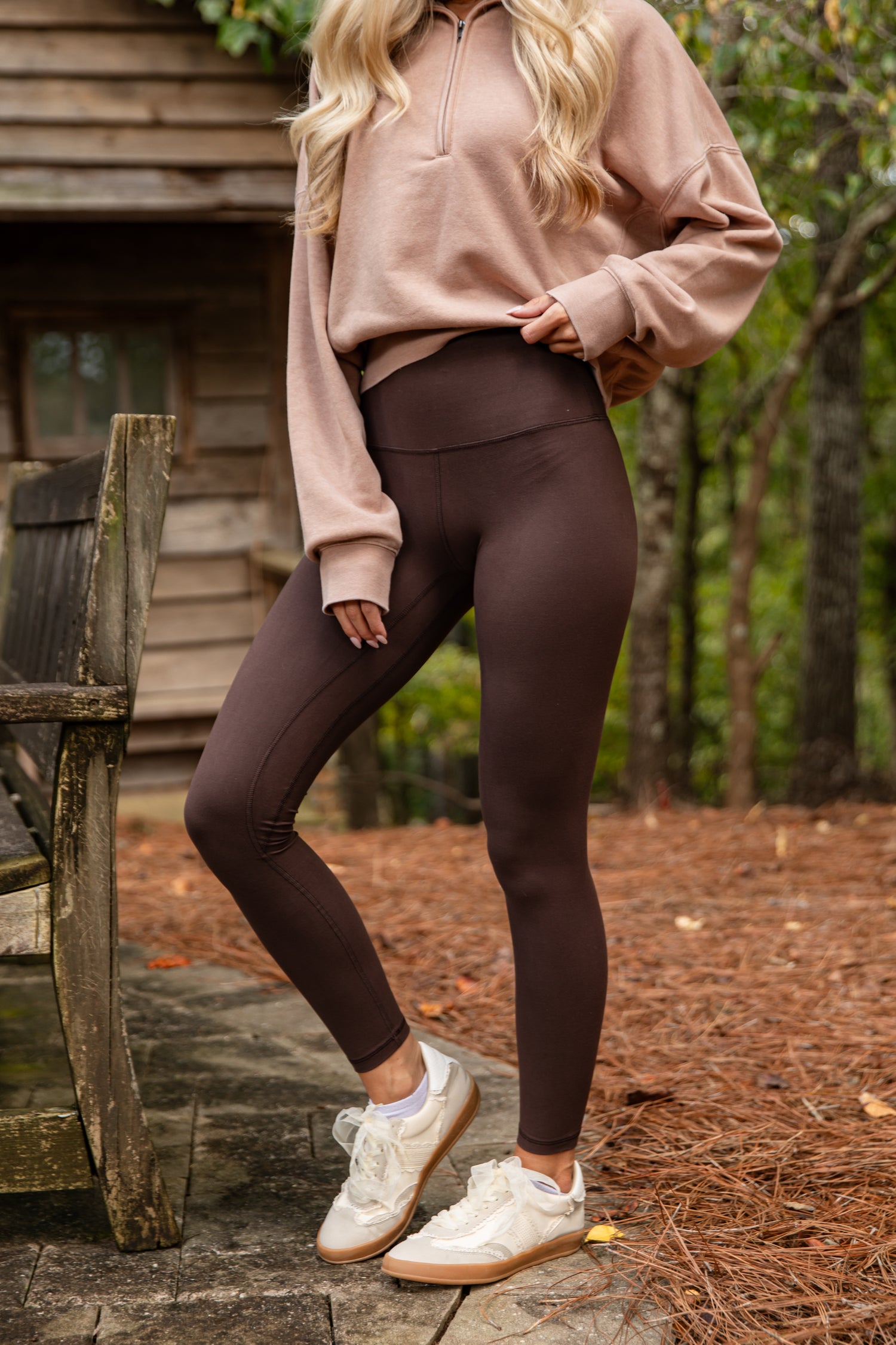 Person wearing a beige sweatshirt and brown leggings standing outdoors near a wooden structure.