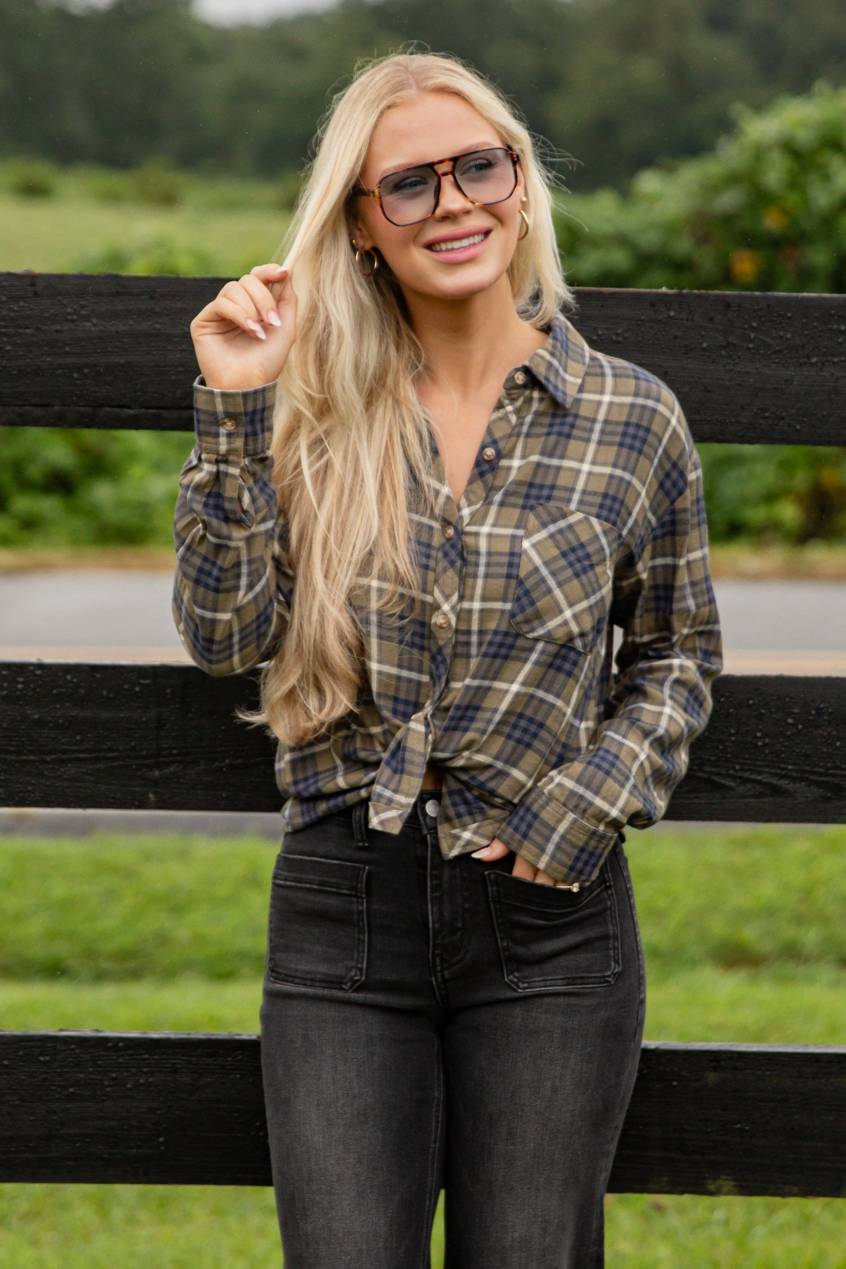 Woman wearing a plaid shirt and sunglasses standing in front of a black fence with greenery in the background
