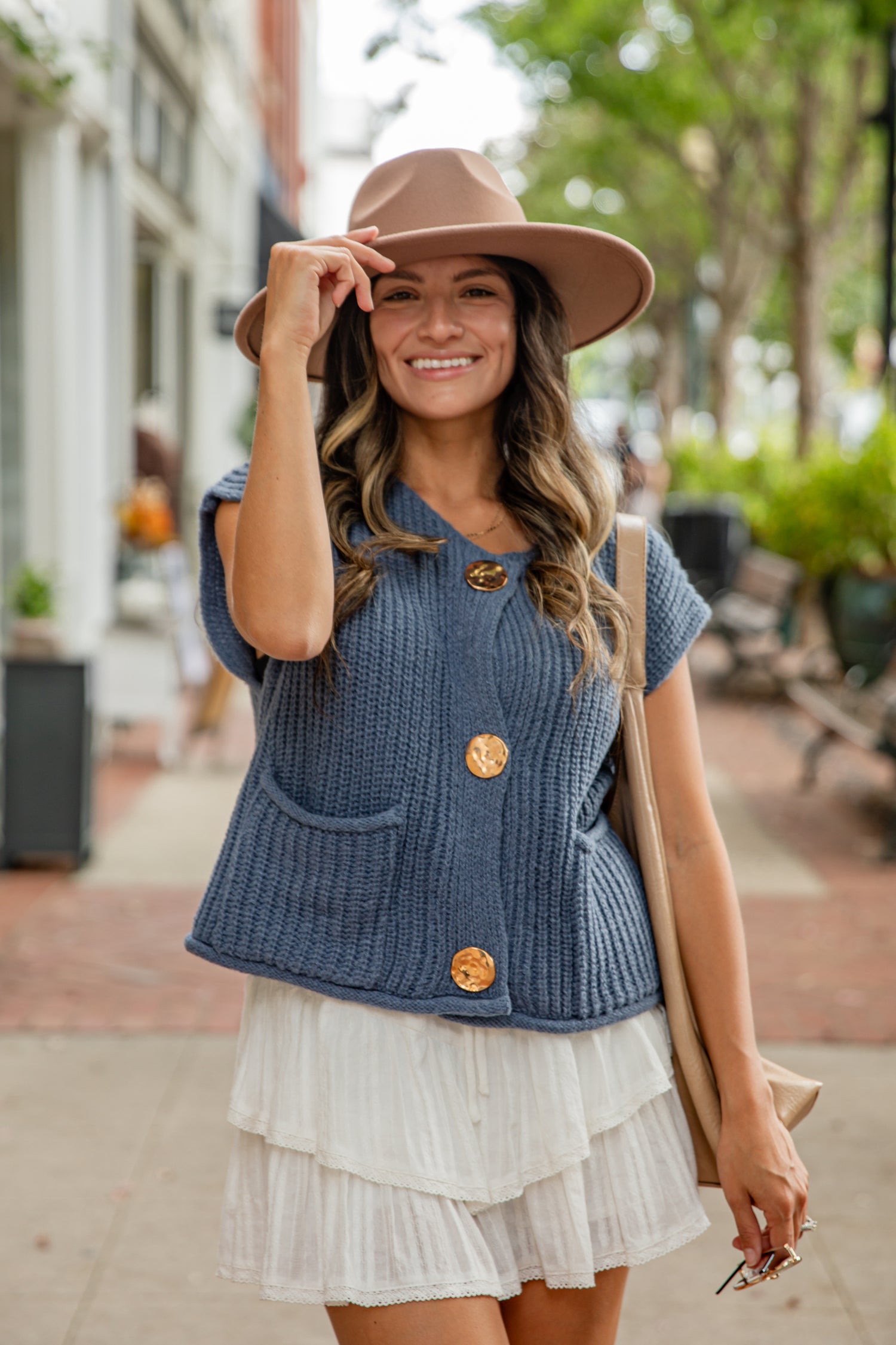 Woman wearing a blue cardigan with gold buttons, white skirt, and brown hat outdoors.