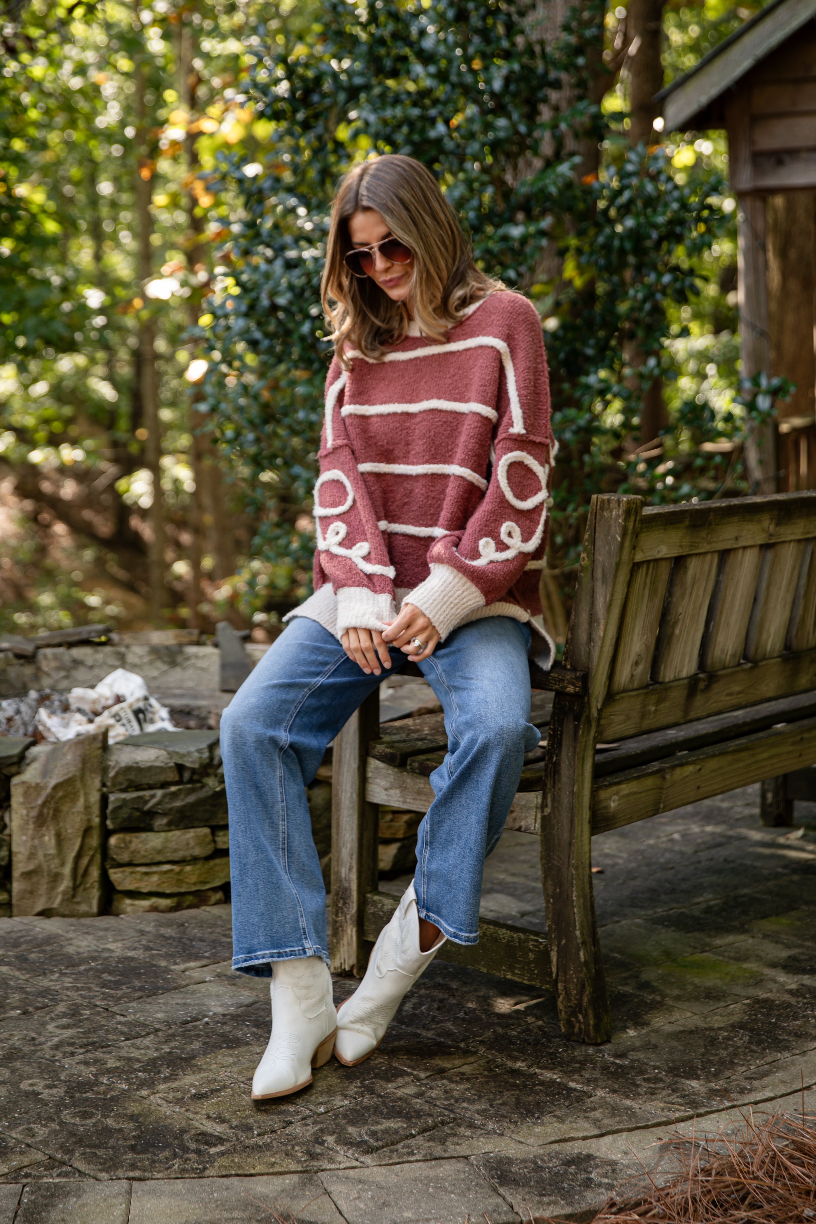 Woman sitting on a wooden bench outdoors wearing a striped sweater, jeans, and white boots.
