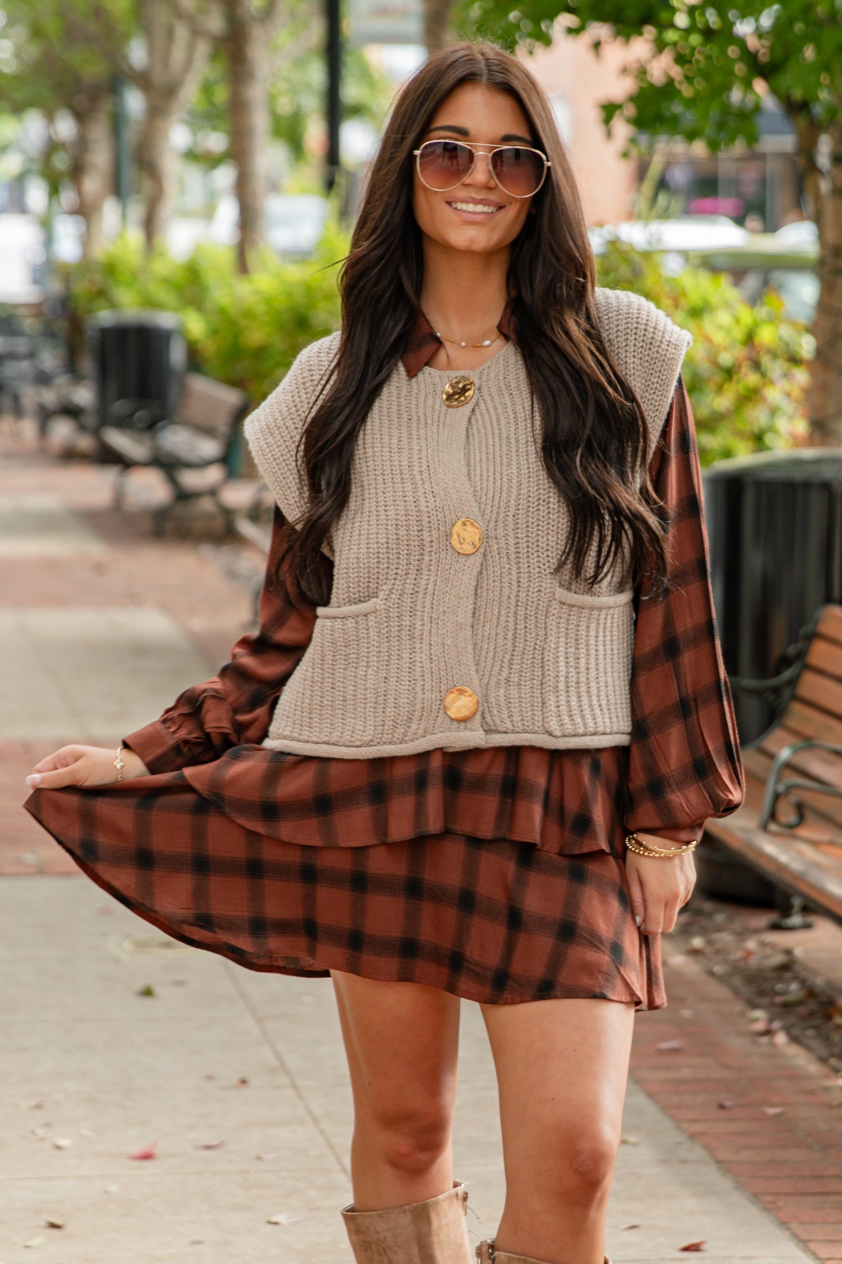 Woman in plaid dress and cardigan standing on a sidewalk with trees and benches in the background