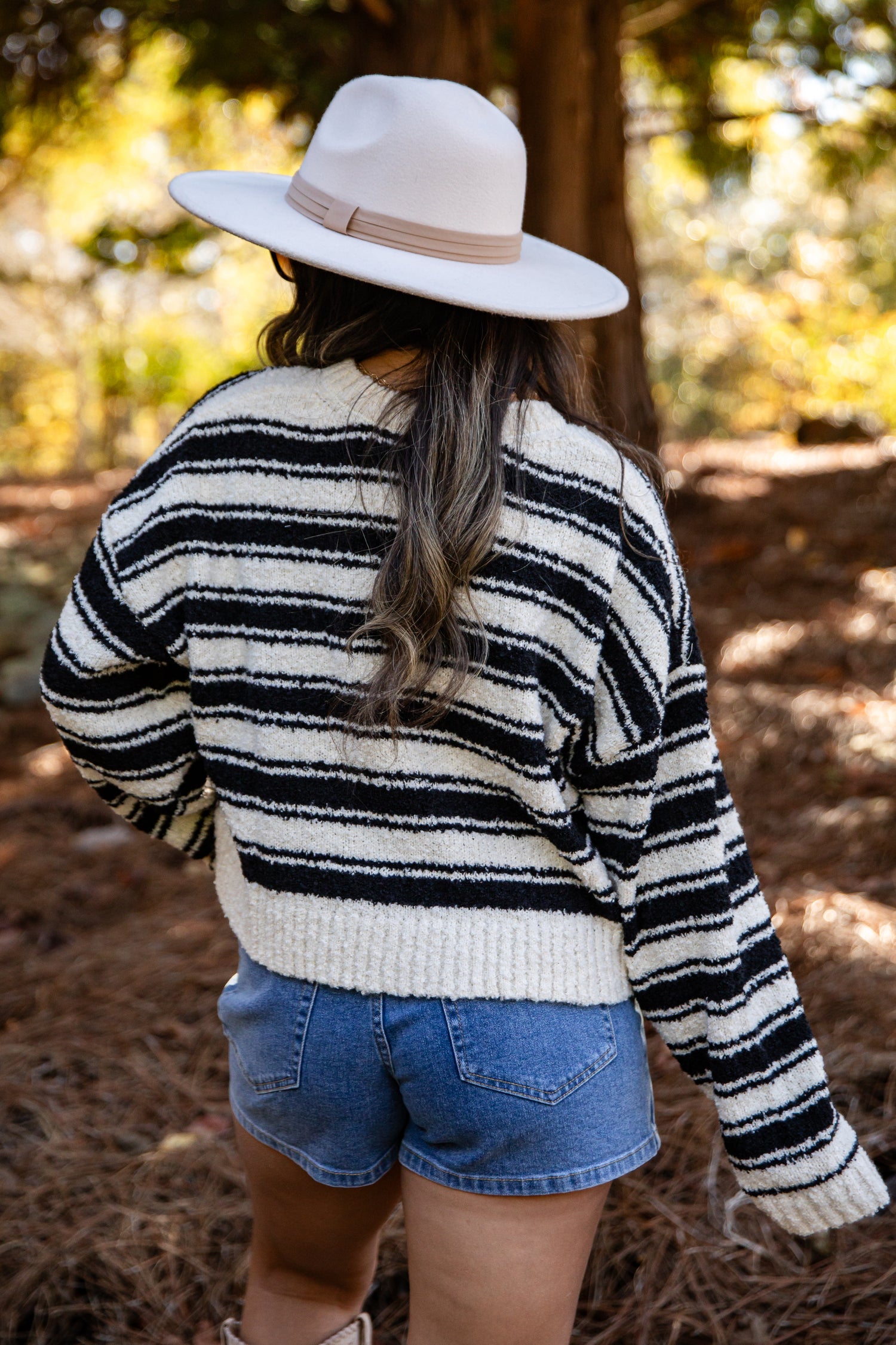 Person wearing a black and white striped sweater and denim shorts with a wide-brimmed hat in a forest setting.