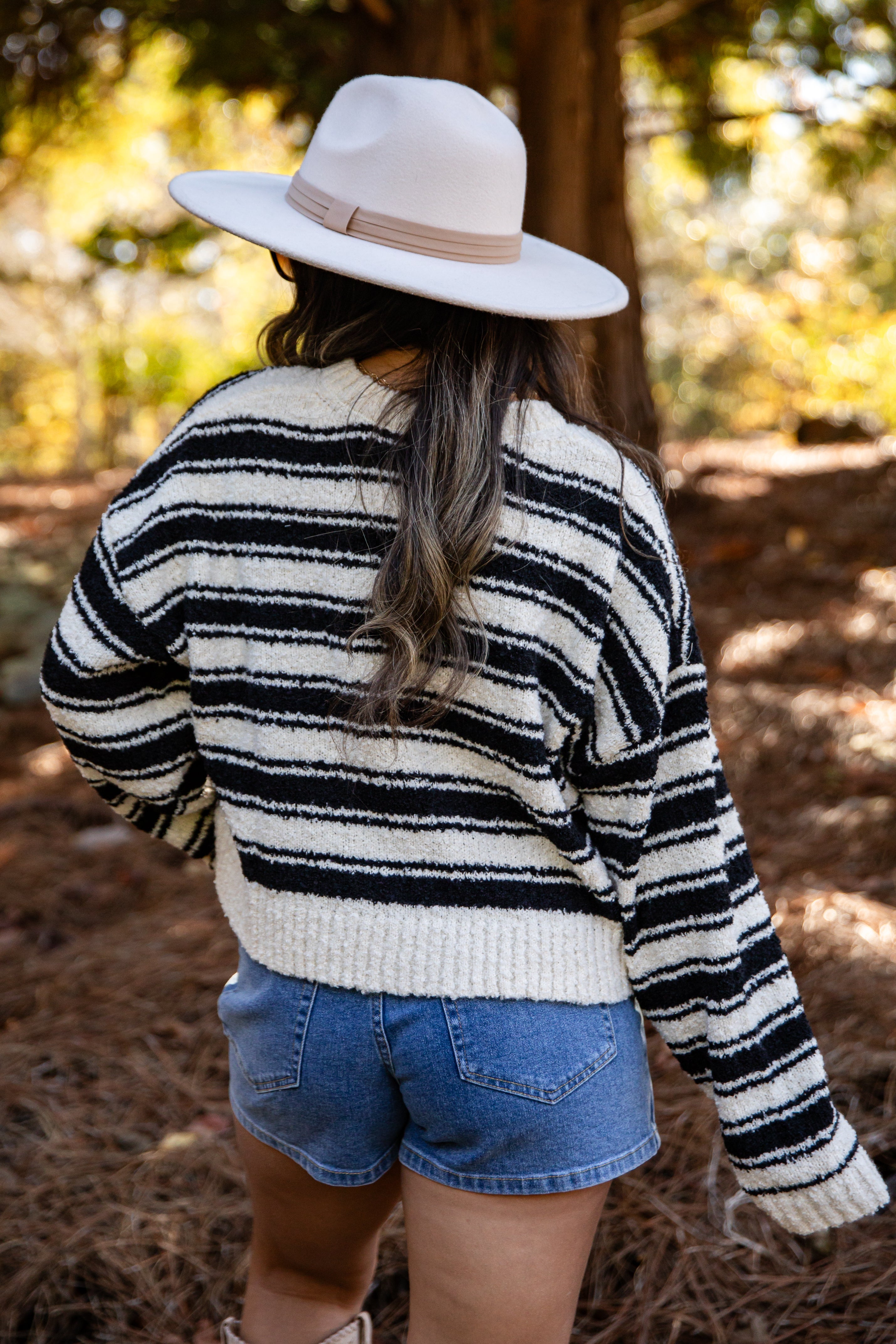 Person wearing a black and white striped sweater and denim shorts with a wide-brimmed hat in a forest setting.