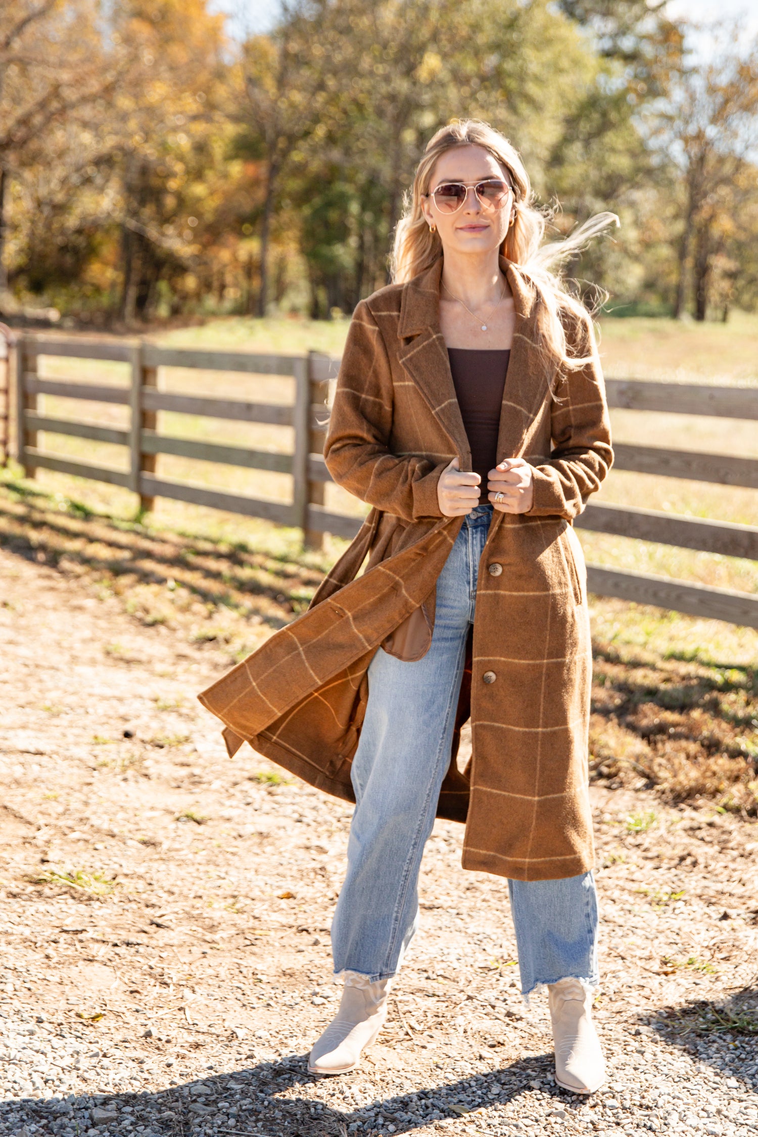 Woman wearing a brown plaid coat in an outdoor setting with trees and a fence.