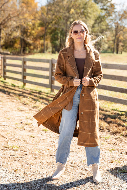 Woman wearing a brown plaid coat in an outdoor setting with trees and a fence.