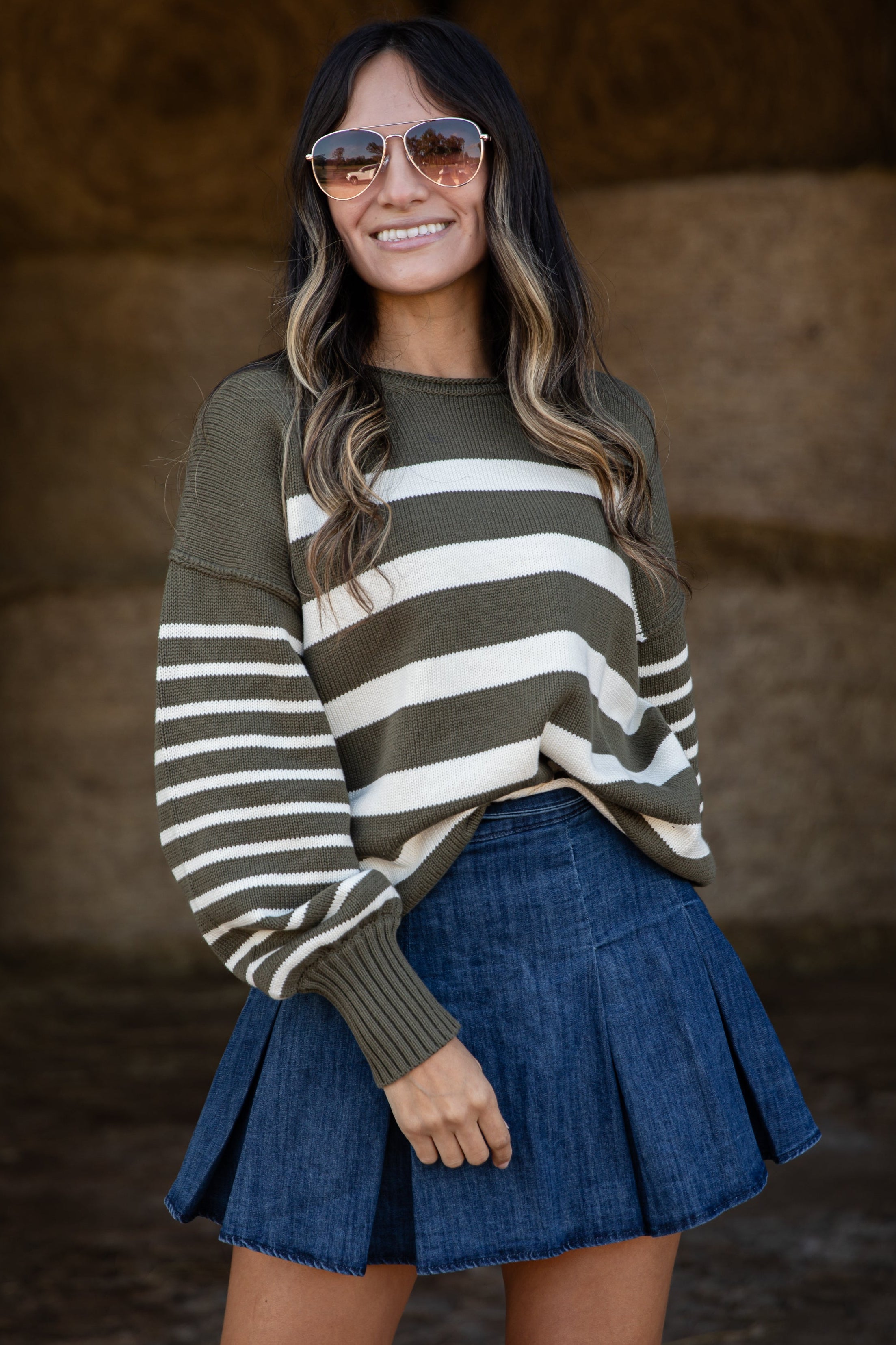 Woman wearing a striped sweater and denim skirt standing in front of hay bales.