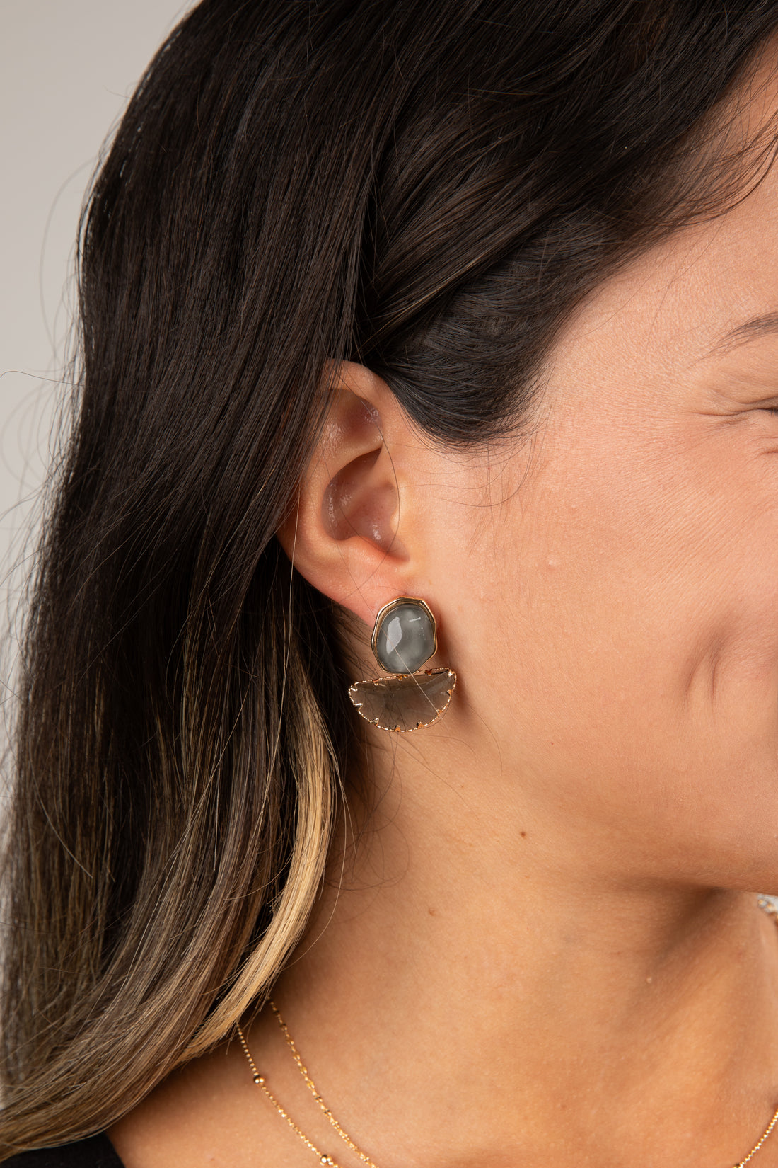 Close-up of a person wearing gold earrings with a gray stone on a neutral background