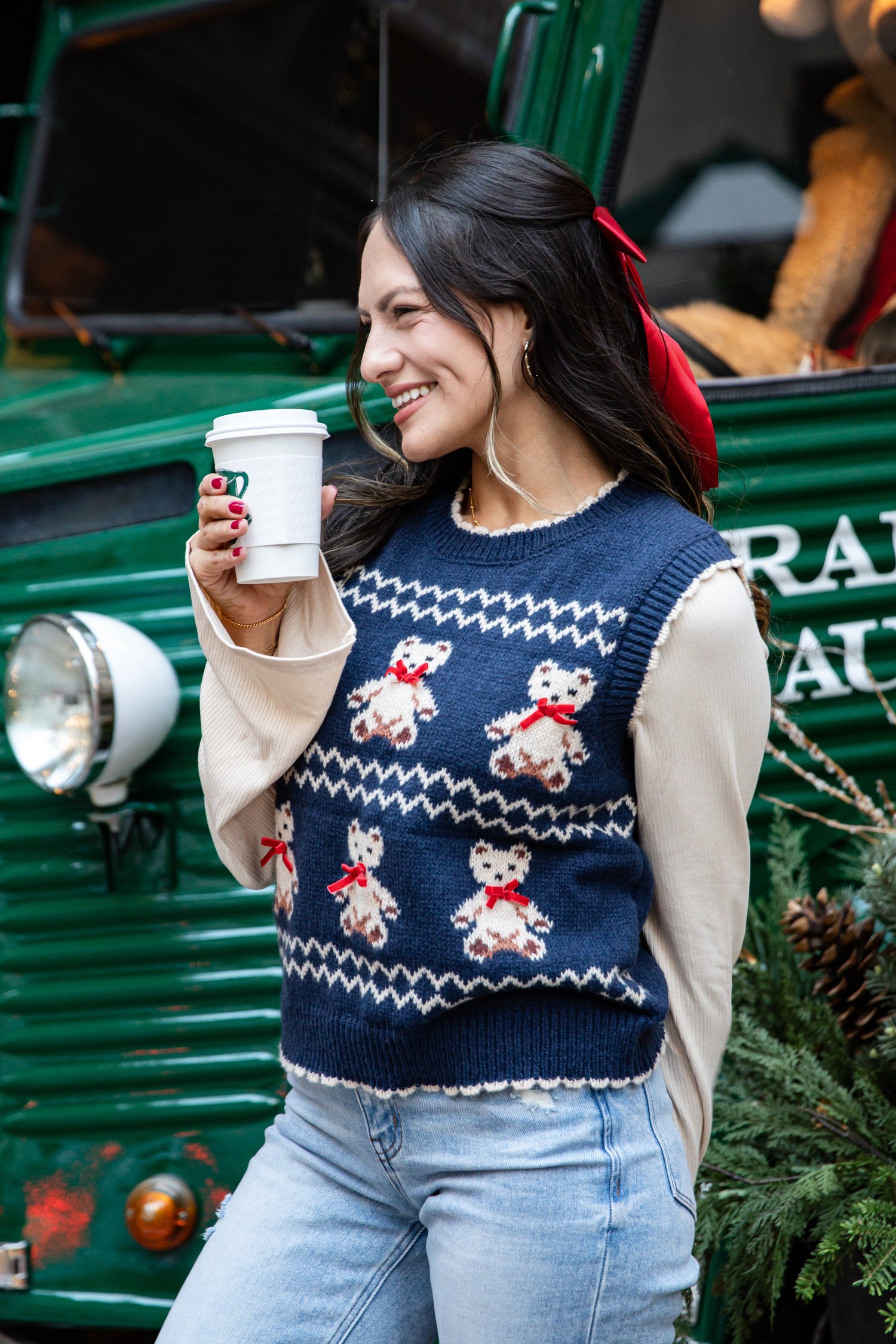Woman wearing a navy sweater with teddy bear designs, holding a coffee cup, standing in front of a green truck.