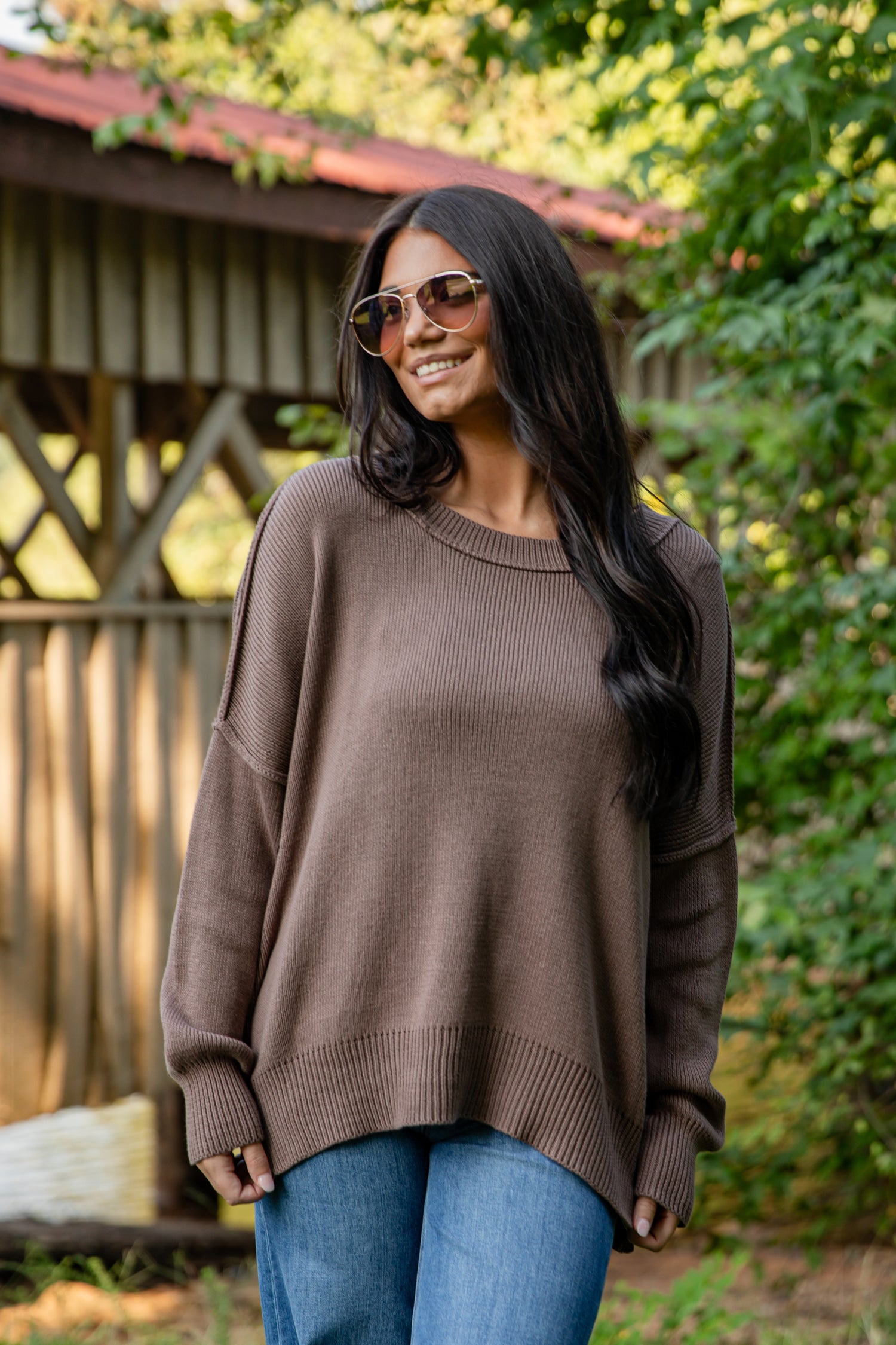 Woman wearing a brown sweater and sunglasses outdoors with greenery in the background