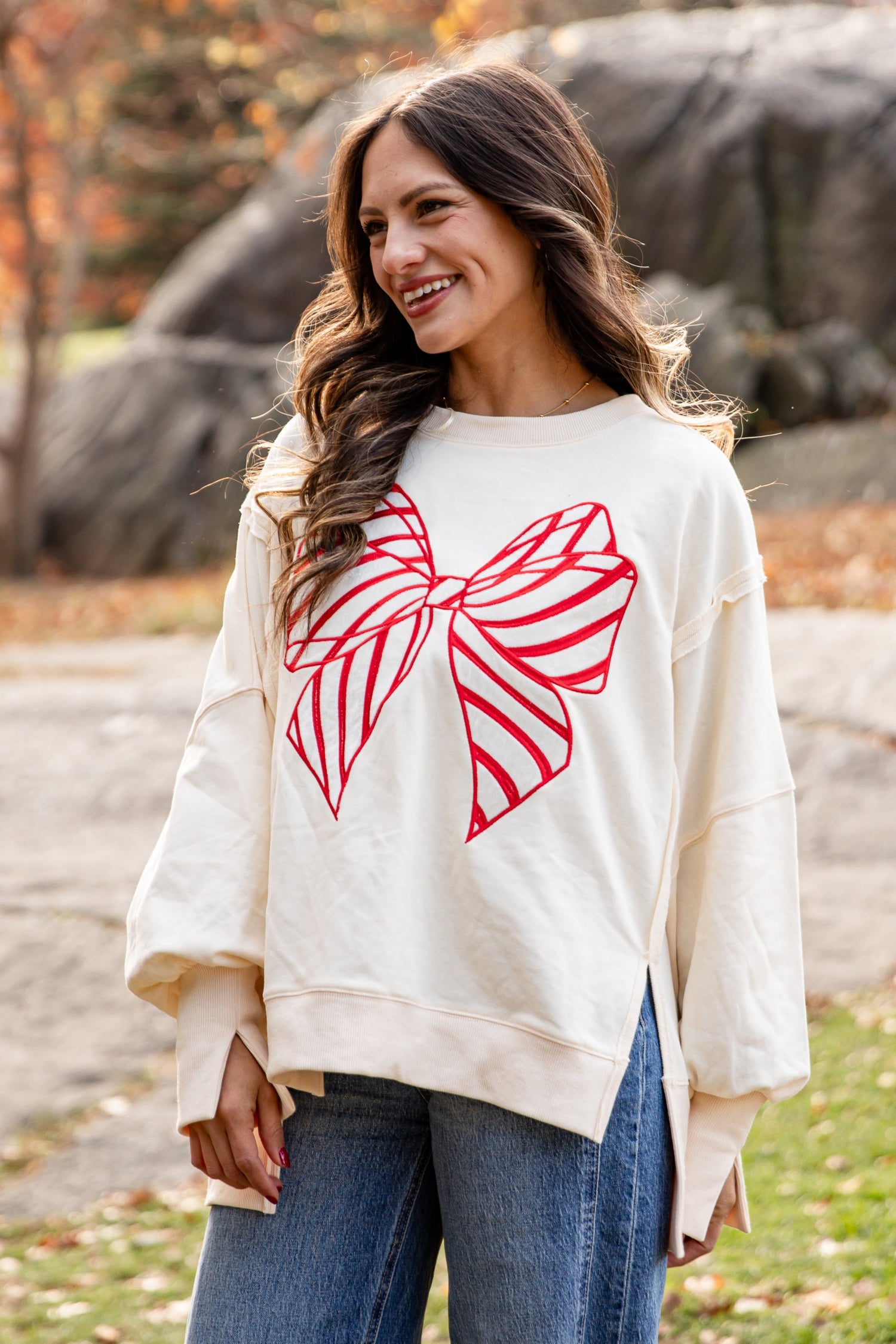 Woman wearing a cream sweater with a red bow design in an outdoor setting with trees and rocks.