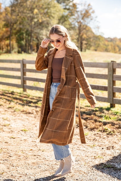 Woman wearing a brown plaid coat in an outdoor setting with trees and a wooden fence.