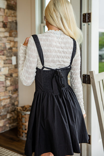 Woman wearing a white blouse and black pinafore dress standing in a doorway.