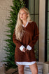Woman wearing a brown sweater over a white shirt in a cozy indoor setting with a Christmas tree.