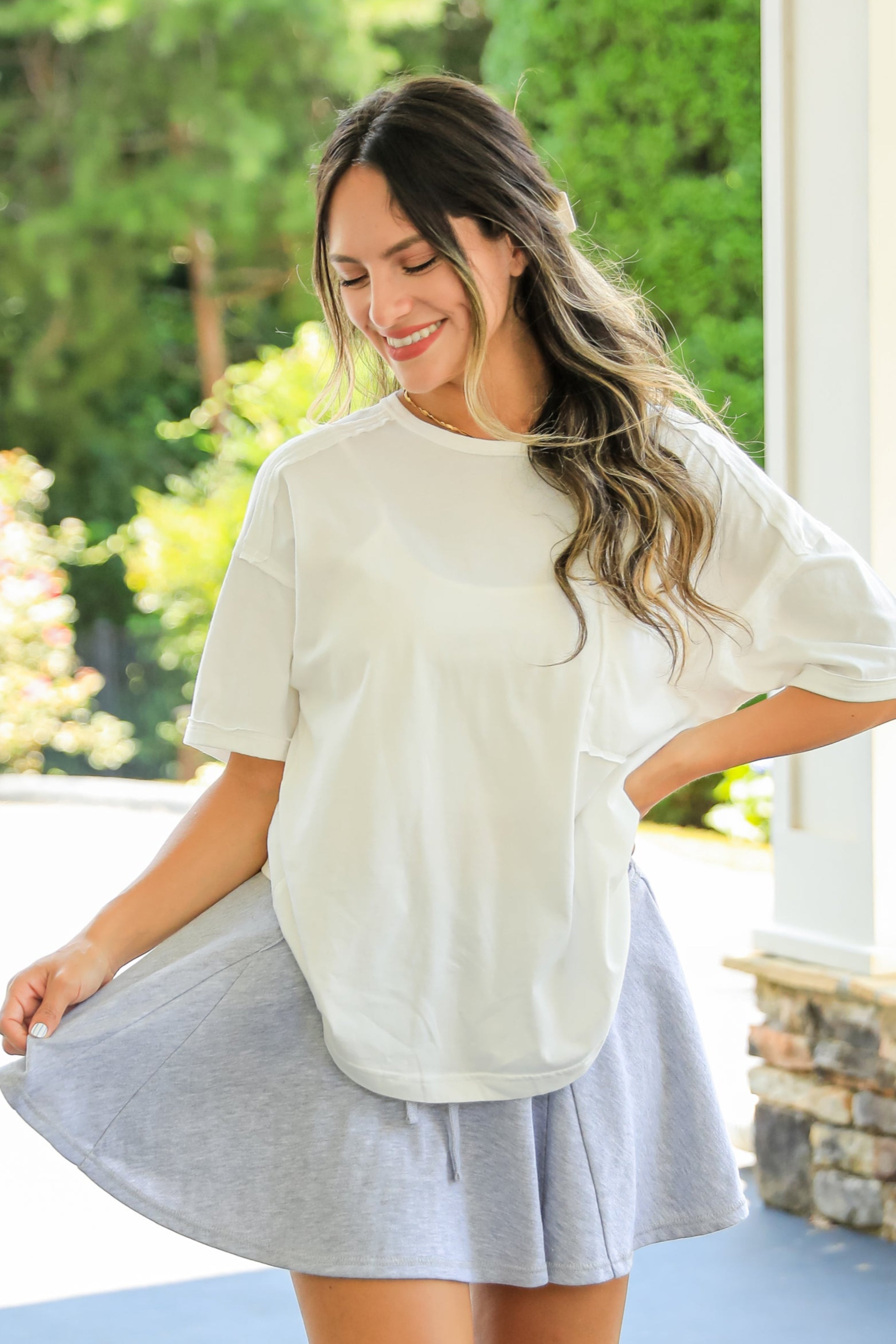 Woman wearing a white oversized shirt and light blue skirt standing on a patio with greenery in the background.