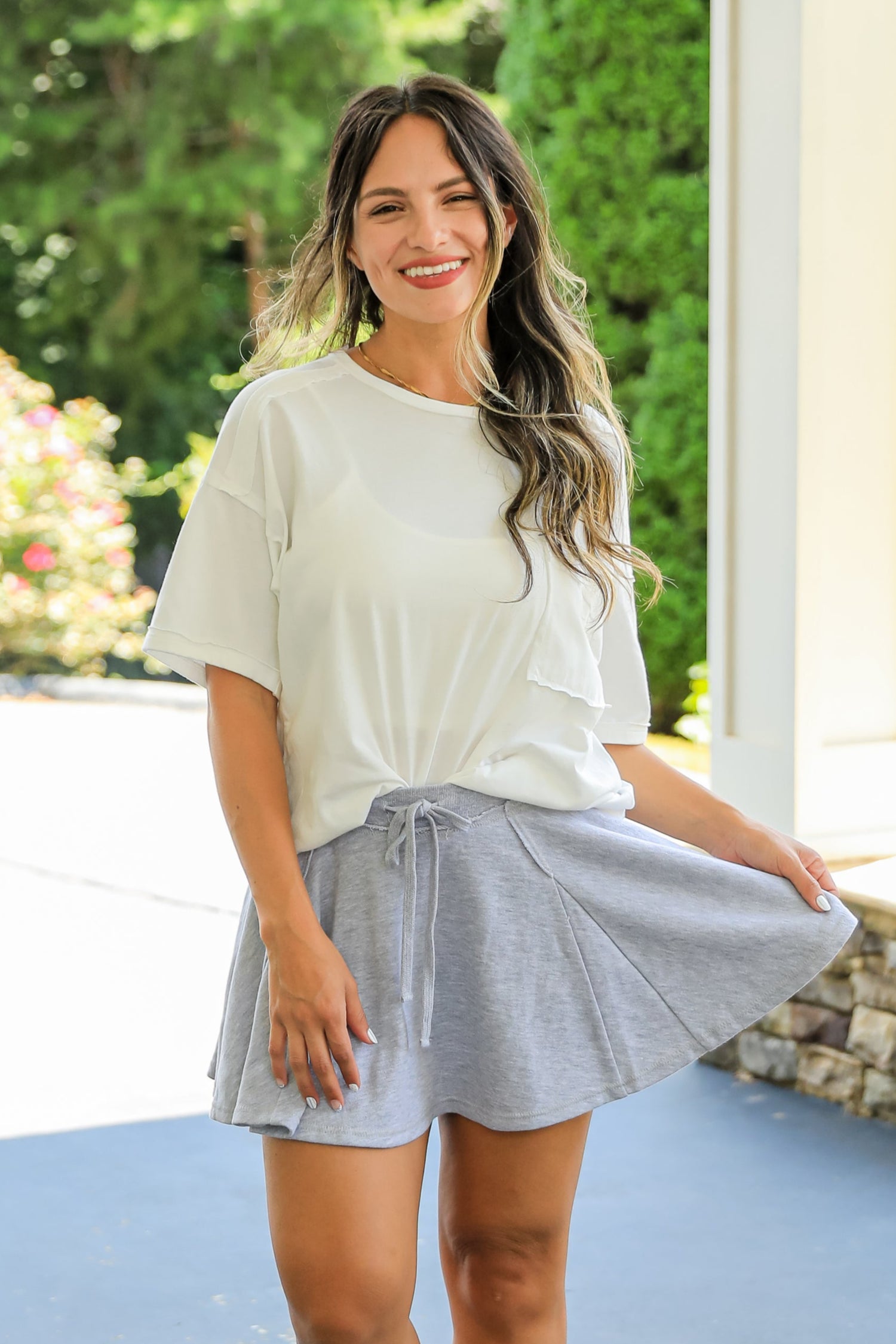 Woman in a white top and gray skirt standing on a patio with greenery in the background