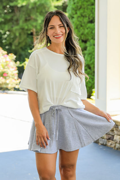 Woman in a white top and gray skirt standing on a patio with greenery in the background