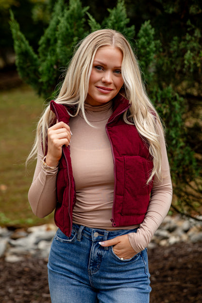 Woman wearing a red vest over a beige top and blue jeans outdoors.