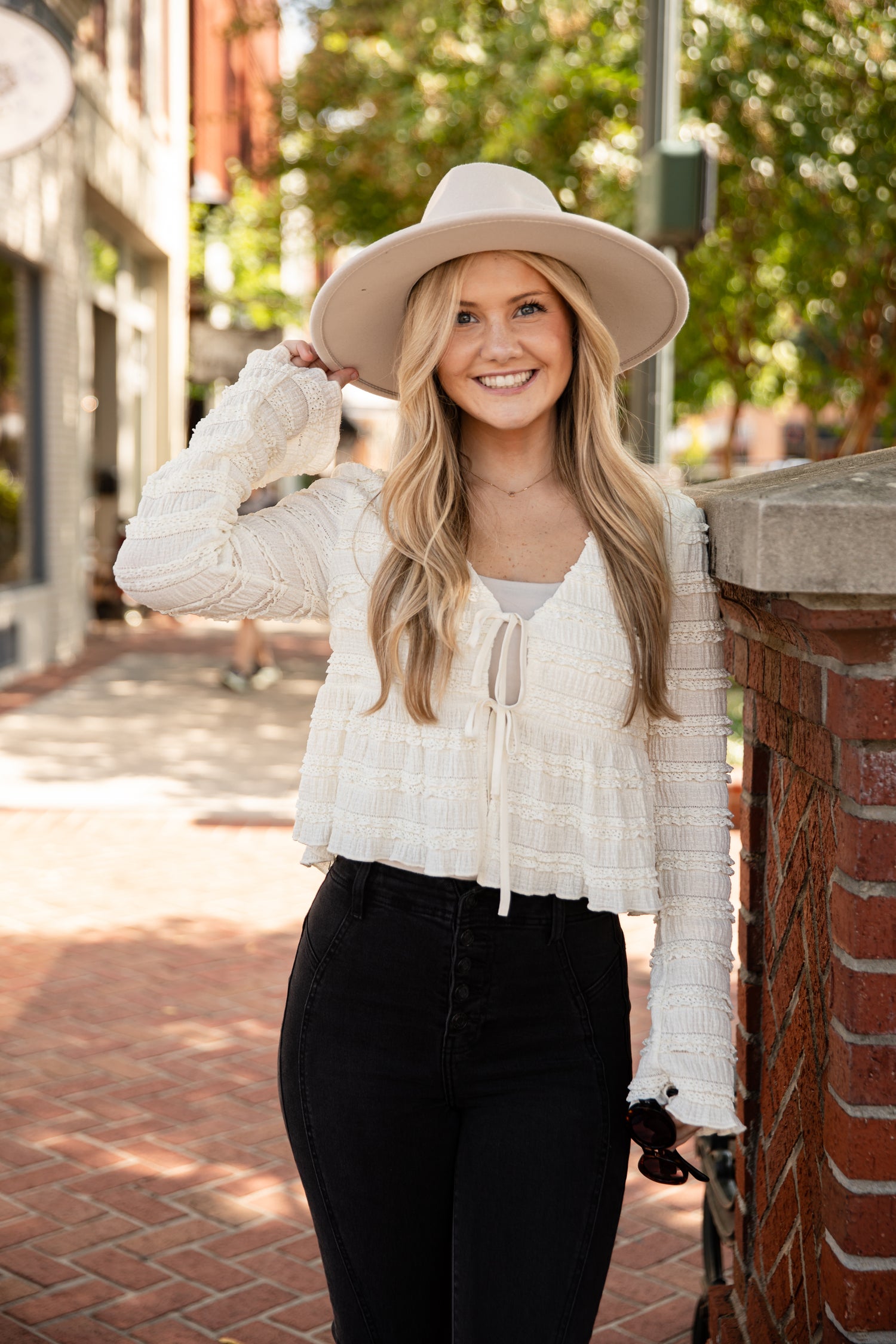 Woman wearing a white lace top and black pants with a beige hat on a city street.