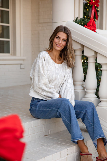 Woman sitting on a staircase wearing a white sweater and blue jeans, with festive decorations in the background.