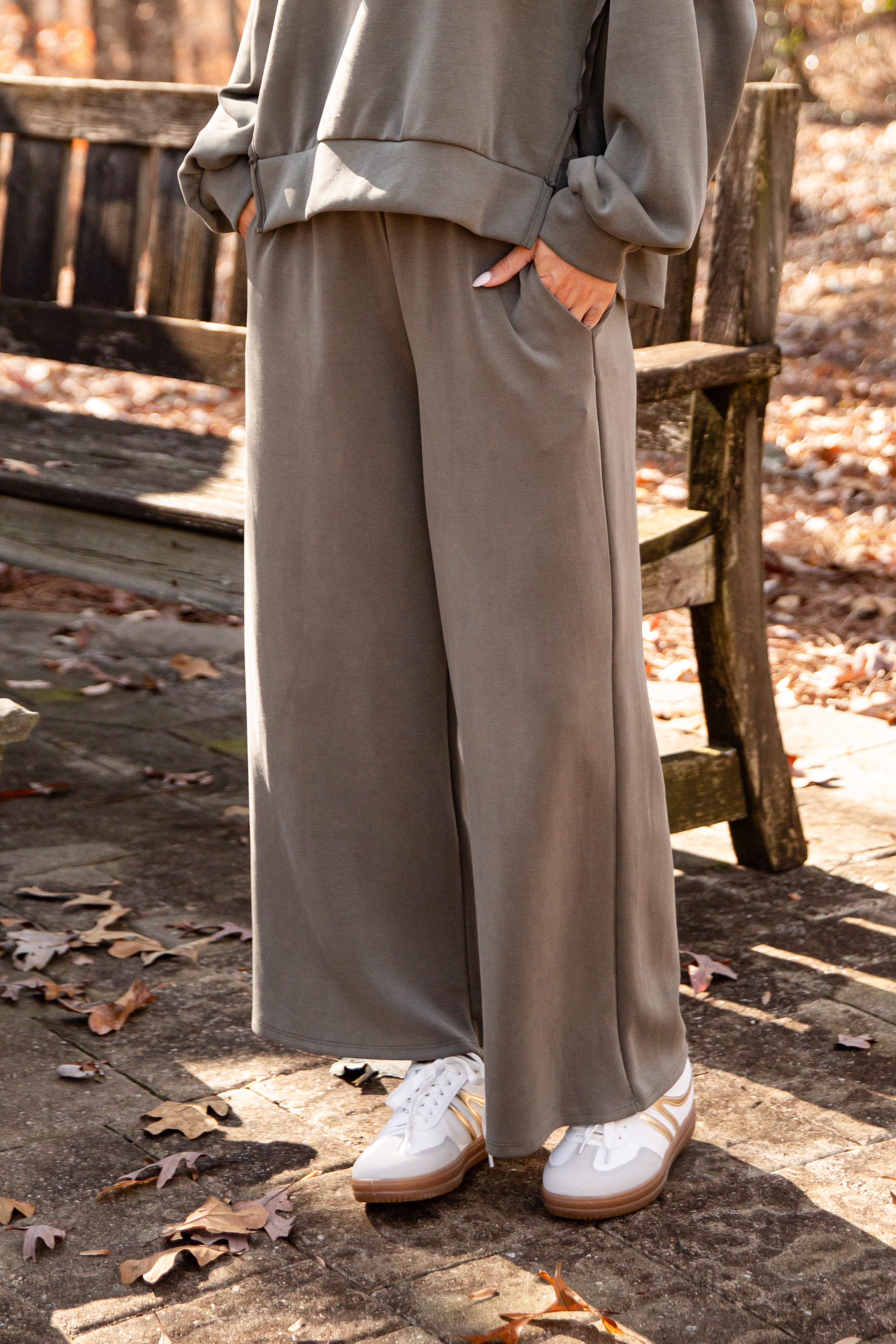 Person wearing a gray outfit with wide-leg pants sitting on a wooden bench outdoors.