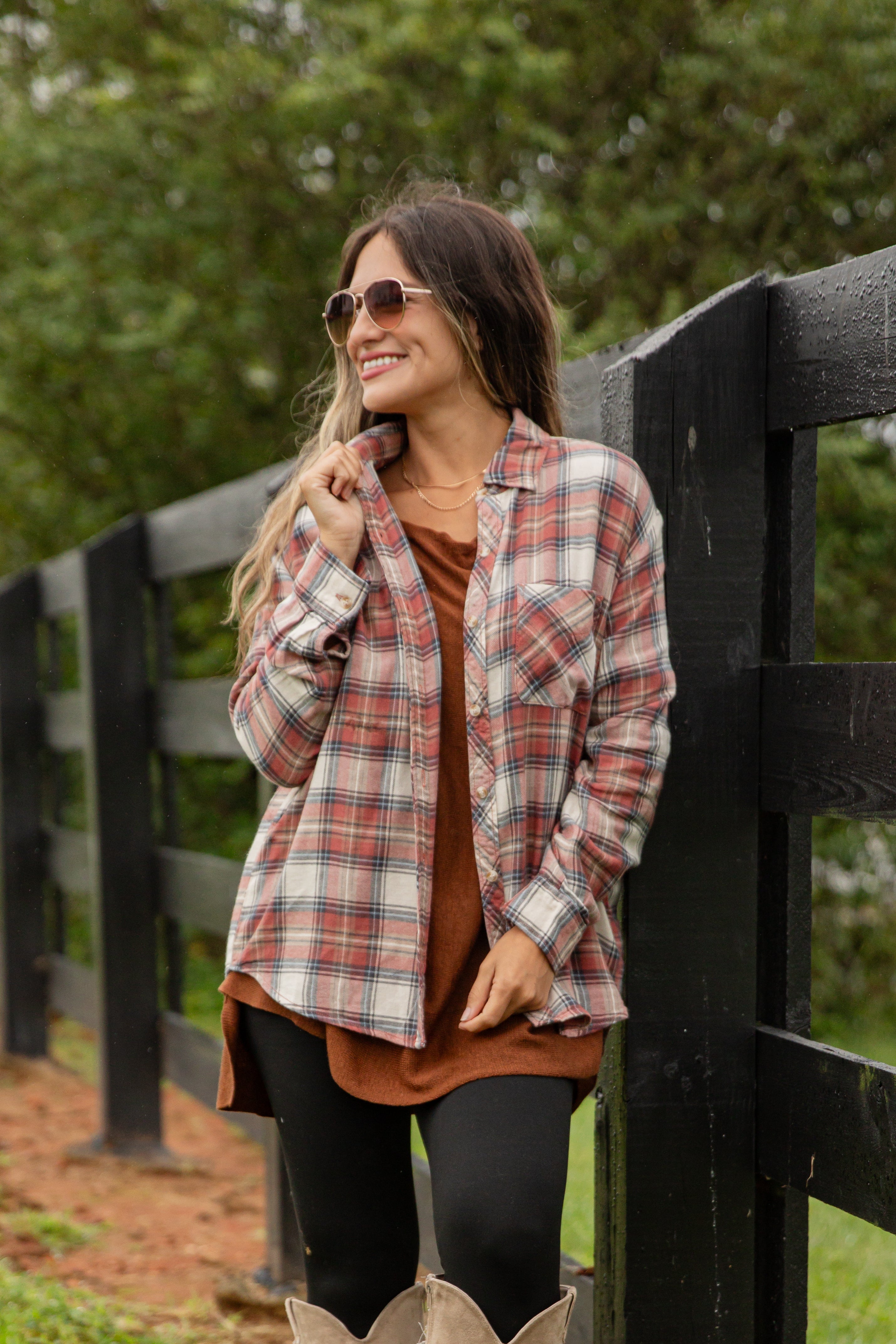 Woman wearing a plaid shirt over a brown top, standing in front of a wooden fence with greenery in the background.