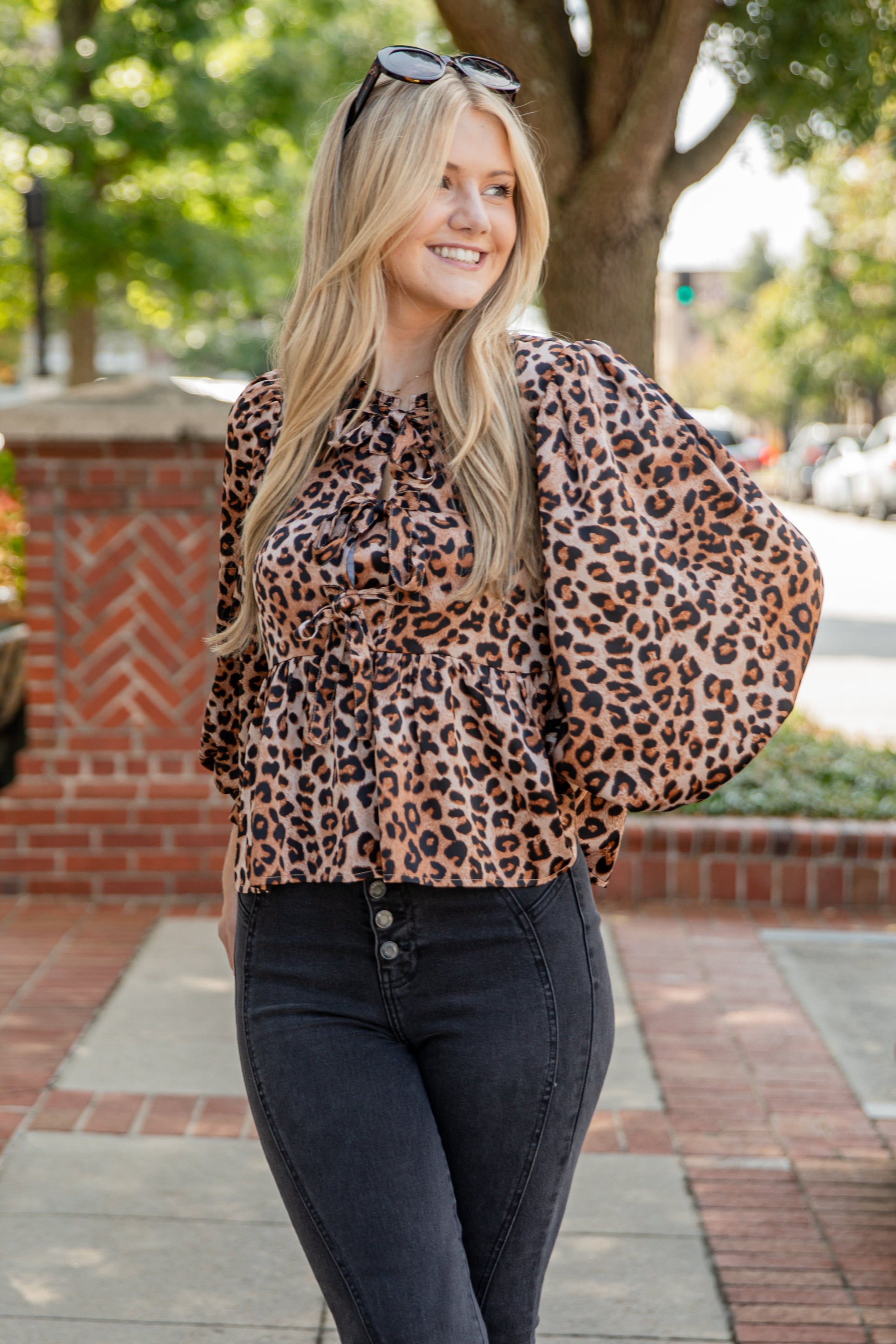 Woman wearing a leopard print blouse and black pants standing outdoors on a sidewalk.