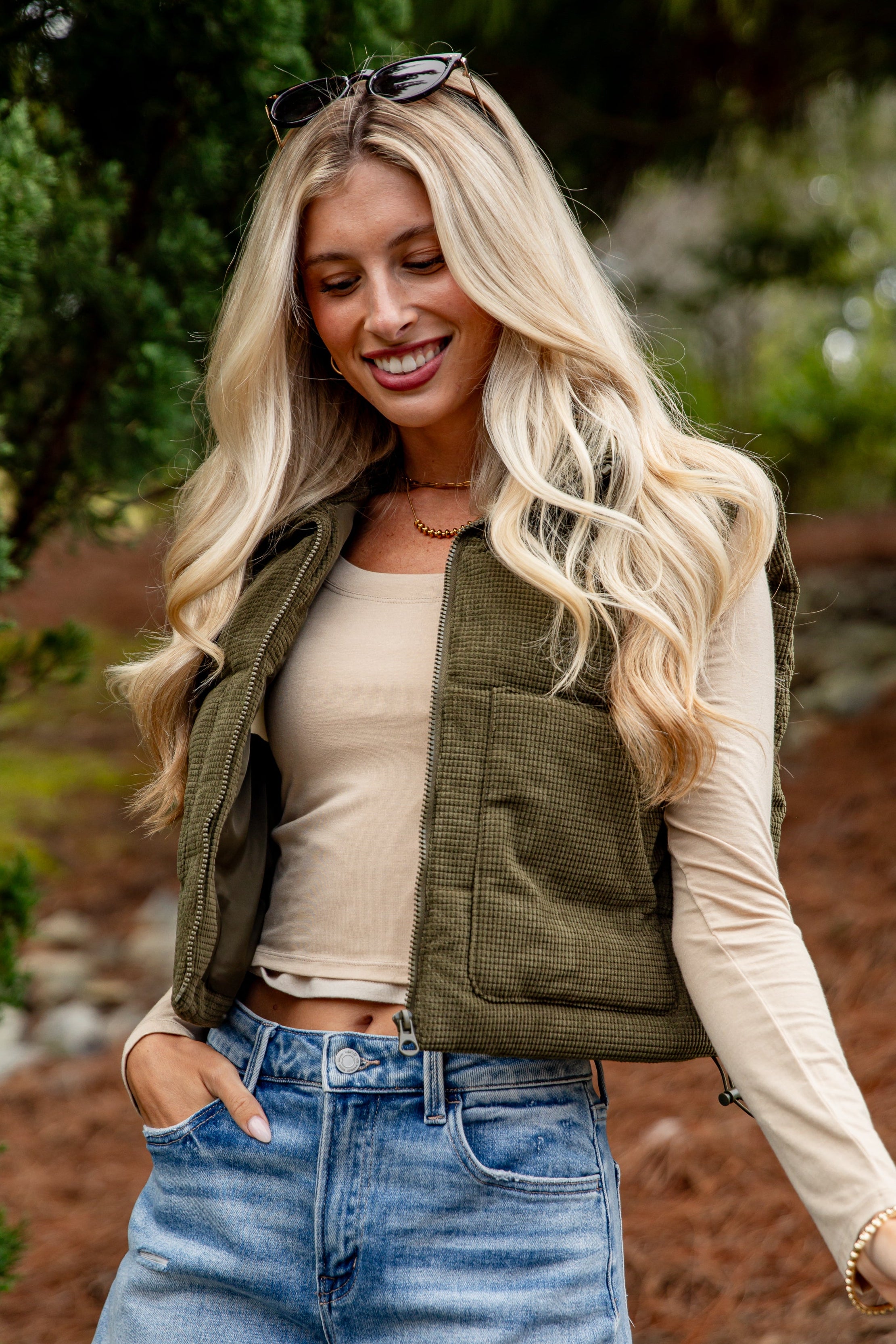 Woman wearing a green vest, beige long-sleeve shirt, and denim shorts outdoors.