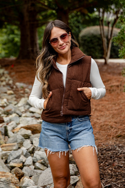 Woman wearing a brown vest, white shirt, and denim shorts outdoors.