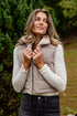 Woman wearing a beige puffer vest and gray jeans standing outdoors with greenery in the background