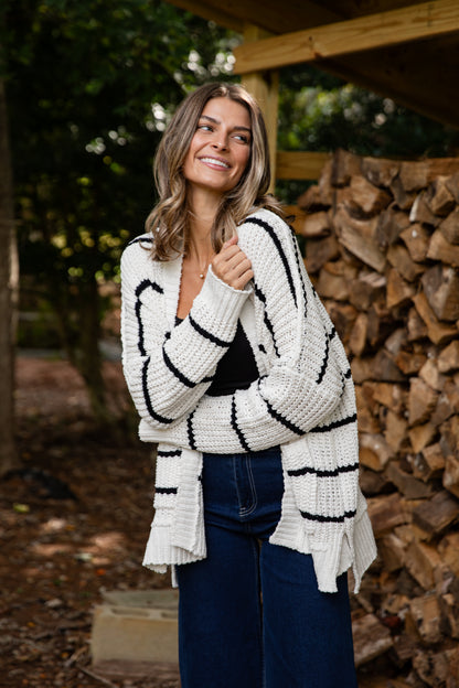 Woman wearing a white cardigan with black stripes standing in front of stacked firewood.