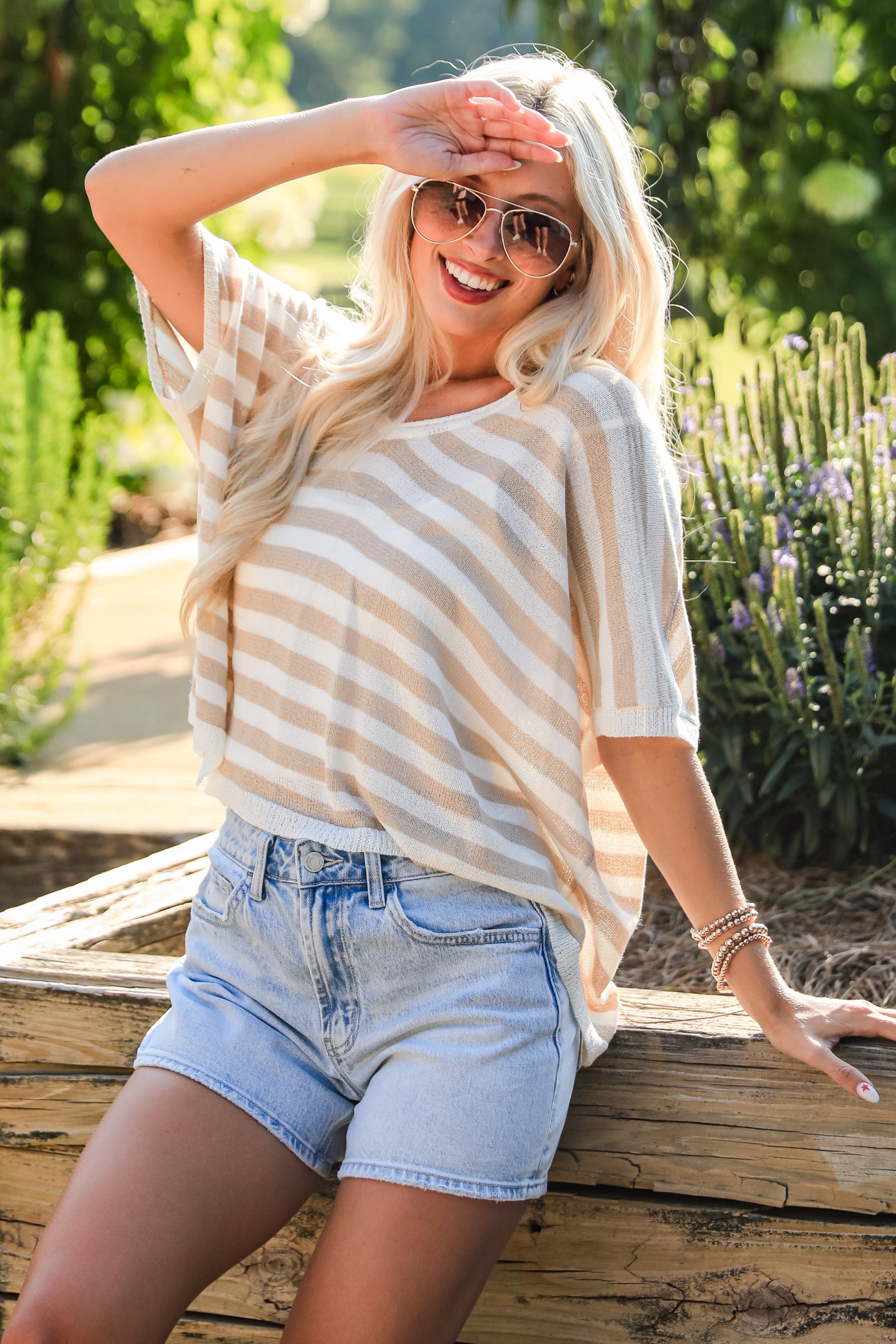 Woman in a striped shirt and denim shorts sitting on a wooden bench outdoors.